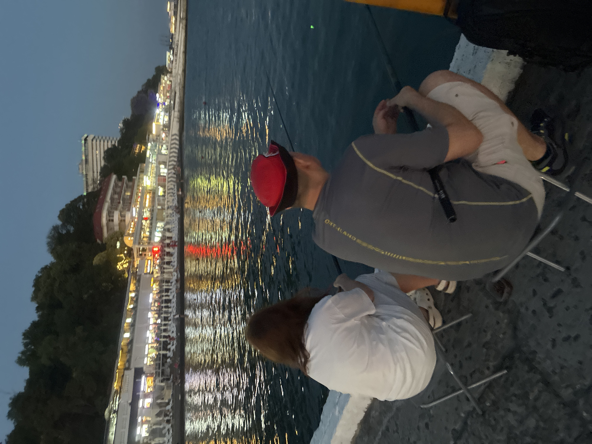 Two people are fishing from a pier in Sochi, Russia, at dusk. The pier is made of concrete and is lined with lounge chairs. In the background, there is a row of illuminated buildings, likely restaurants and shops, along the coastline. The water reflects the lights from the buildings. The person in the foreground on the left is wearing a white t-shirt and light-colored shorts, and the person on the right is wearing a gray t-shirt, light-colored shorts, and a red hat. Both are seated on folding chairs and holding fishing rods. The sky is a deep blue, indicating it is twilight.