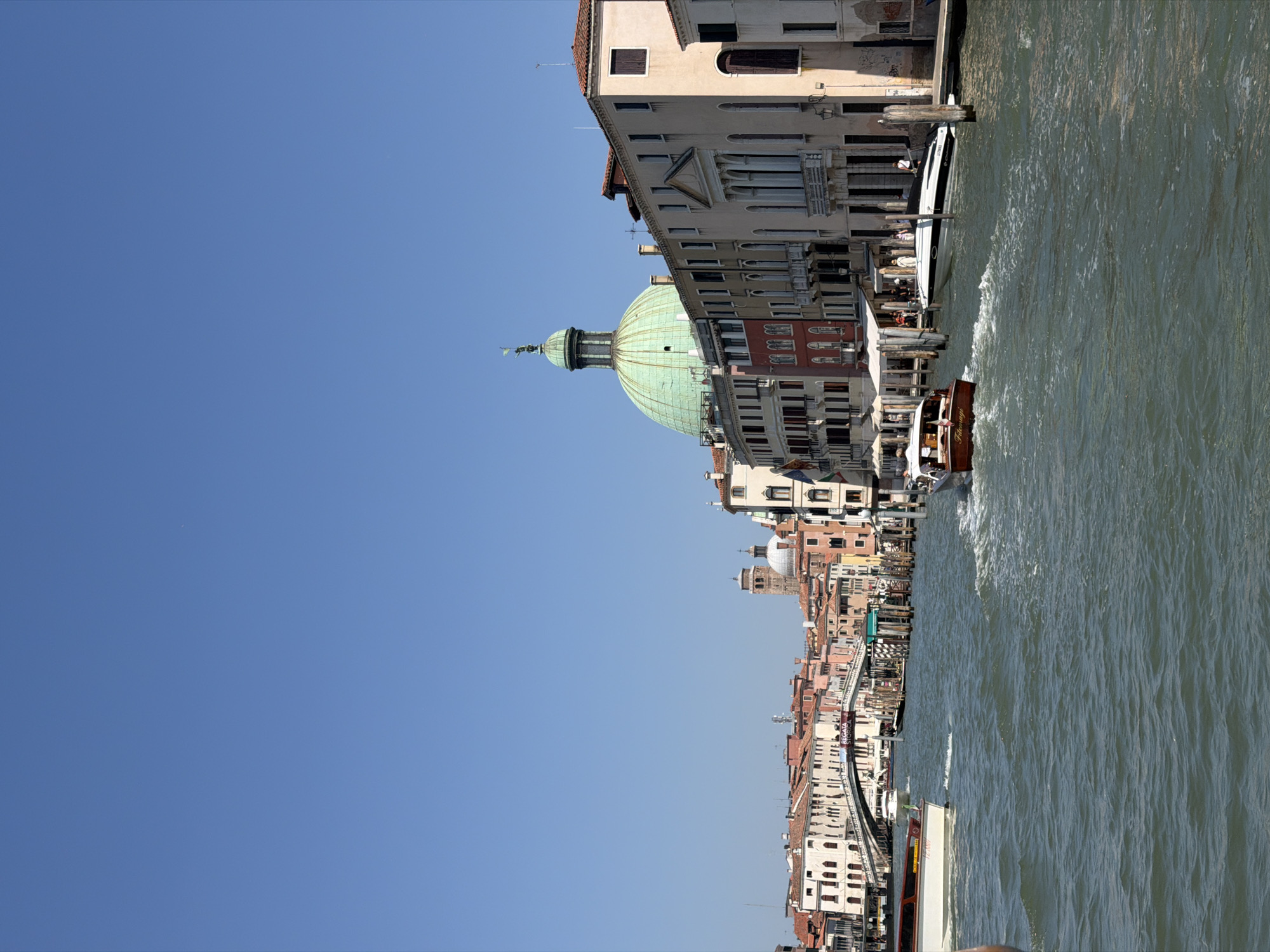 The image depicts a sunny day in Venice, Italy, likely on the Grand Canal. The scene is dominated by a clear blue sky and a body of water with moderate waves. Along the right bank of the canal, a line of historic Venetian buildings with red-tiled roofs and multi-story facades is visible. The architecture includes various styles, with some buildings featuring balconies, arched windows, and decorative elements. Prominent among these buildings is a structure topped with a large, green dome, characteristic of Venetian churches.
Several boats are present on the water. In the foreground, a white boat with a red stripe is moving from left to right. Further into the canal, another boat, possibly a water taxi, is also in motion. In the background, a bridge arches over the canal, connecting the buildings on either side. More boats are visible under and around the bridge, suggesting ongoing canal traffic. People can be seen on the decks of some of the boats and along the canal banks, engaged in various activities. The overall atmosphere is one of a bustling and picturesque urban waterway.