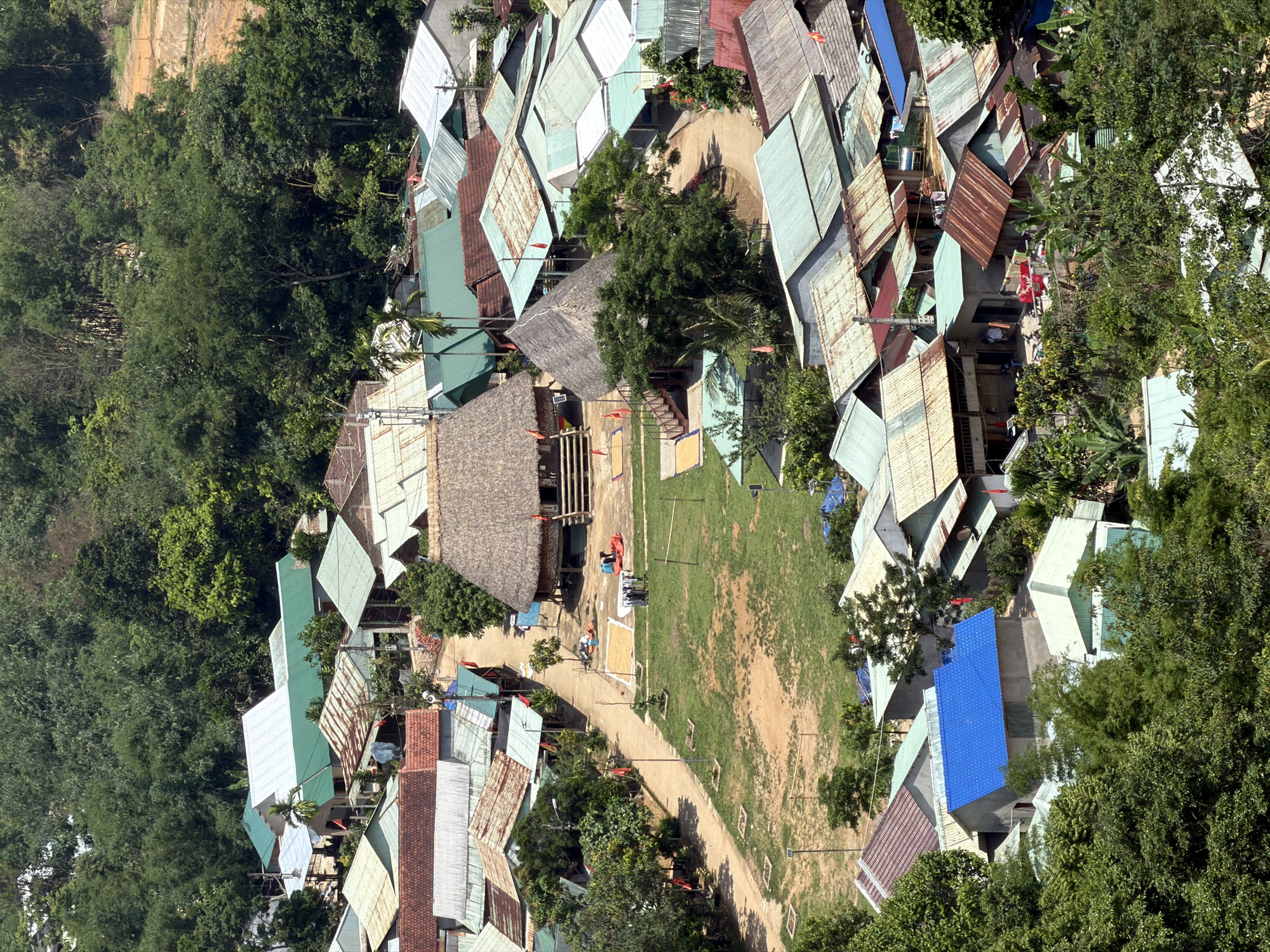 The image shows an aerial view of a settlement within the city of Đà Nẵng, Vietnam. The settlement consists of houses with corrugated metal roofs of varying colors and conditions, interspersed with lush greenery, and clustered around a central clearing with a long thatched-roof structure. A narrow path runs through the settlement and across the clearing. People and objects can be seen within the clearing, possibly related to daily activities. The environment is a mix of dense foliage and built structures.