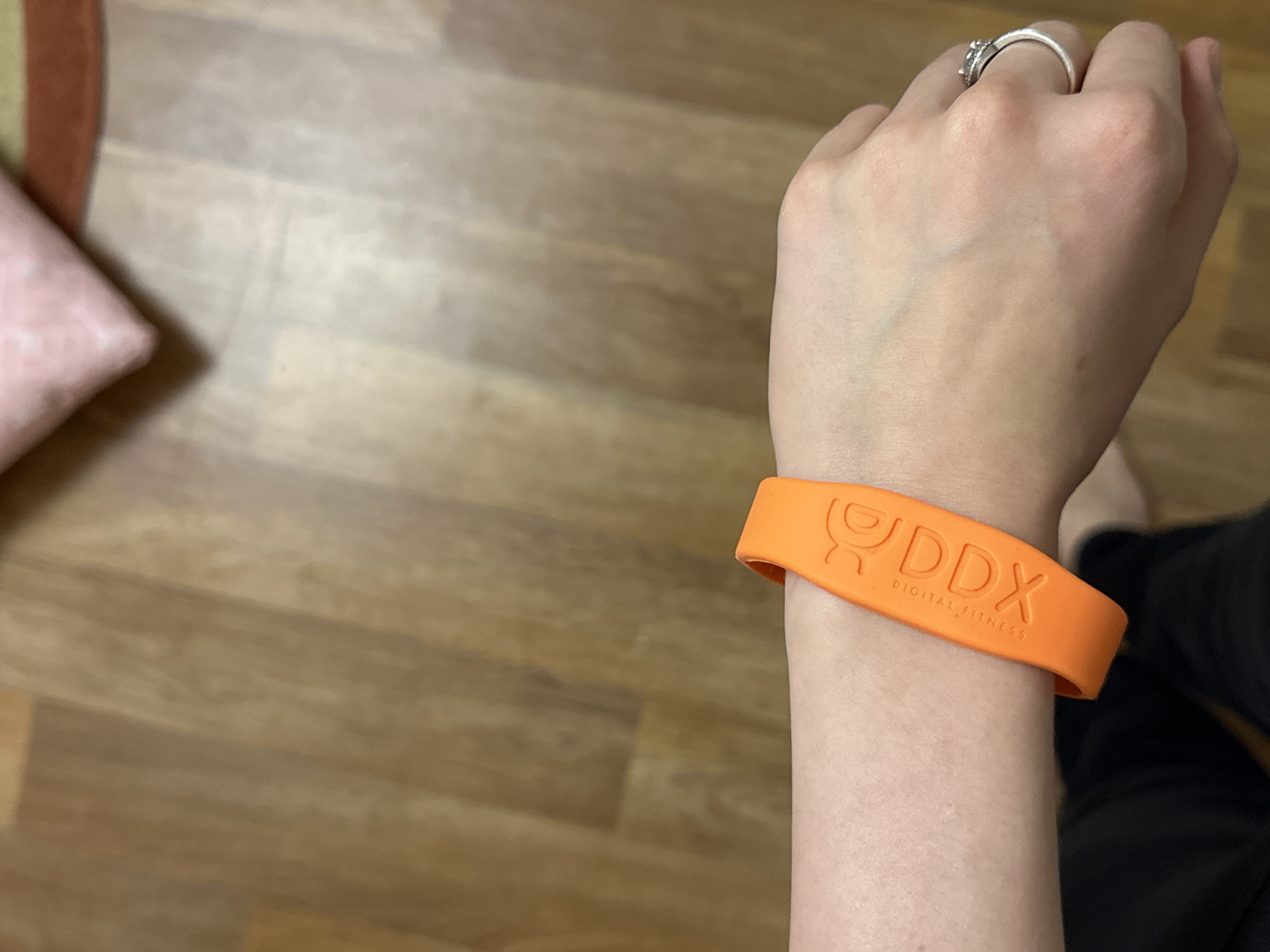 A close-up, eye-level shot shows a person's left arm and hand wearing an orange wristband. The wristband has "DDX" embossed on it in large, stylized letters, with "DIGITAL FITNESS" in smaller text below. The person is also wearing a silver ring with stones on their ring finger. The background consists of light brown wooden flooring, with a hint of a pink and beige patterned cushion visible in the upper right corner. There is no indication of the city of Kazan, Russia in the image.