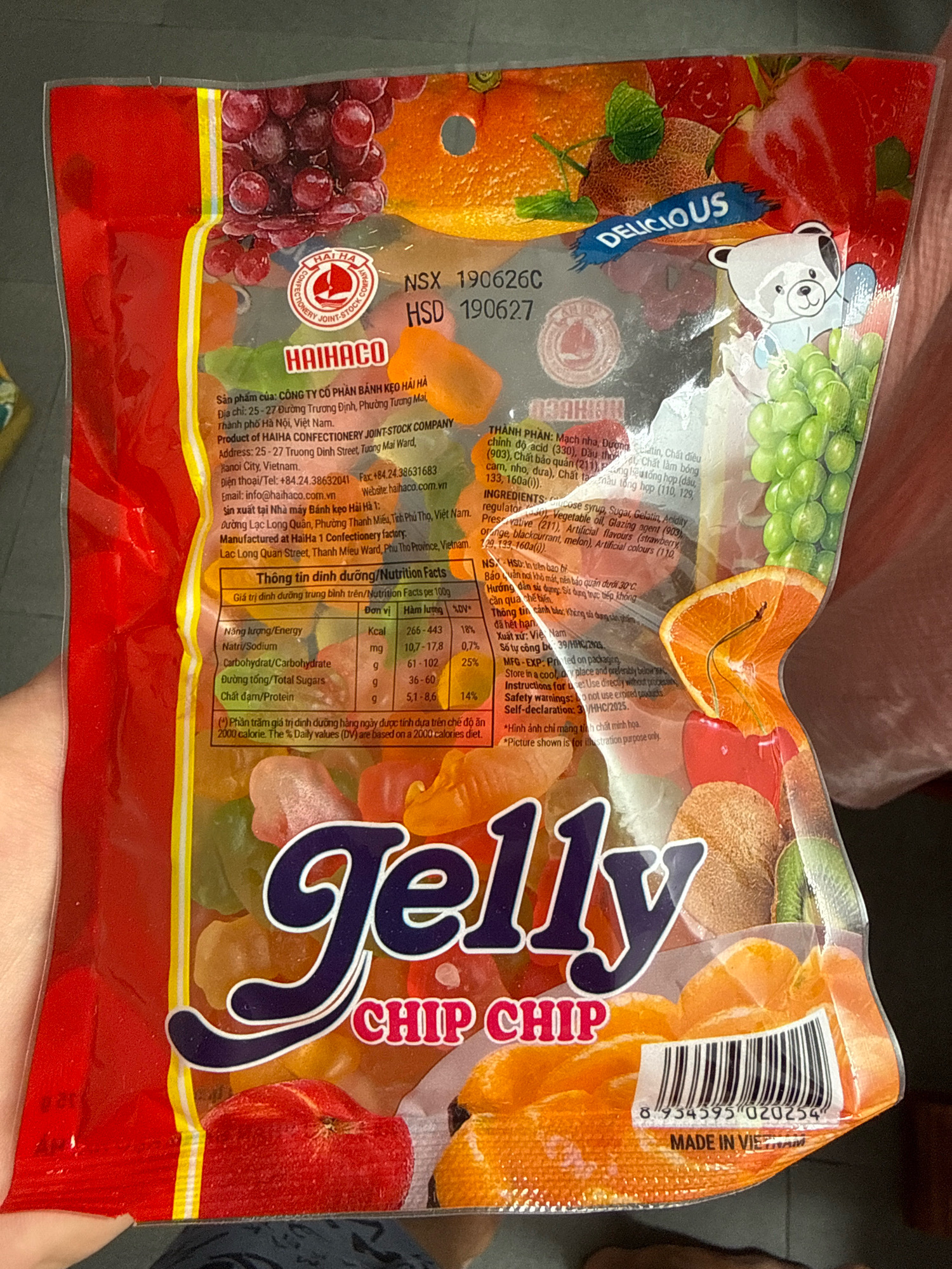 The image is a close-up, top-down shot of a packaged bag of "Jelly Chip Chip" candy. The bag is predominantly red with colorful images of fruits such as grapes, oranges, and strawberries. The text on the bag is in Vietnamese and English, indicating the brand name "HAIHACO" and nutritional information. The product is manufactured by "CÔNG TY CỔ PHẦN BÁNH KẸO HẢI HÀ" (Haiha Confectionery Joint-Stock Company) located at 25-27 Truong Dinh Street, Phuong Mai Ward, Hanoi City, Vietnam. The bag also displays a barcode and the text "MADE IN VIETNAM". The candy inside the bag appears to be gummy candies in various fruit shapes and colors. A person's hand is visible on the left side of the image, holding the bag. The background is dark and out of focus. There is no visible indication of the location being Nha Trang, Vietnam; it is stated that the company is located in Hanoi City, Vietnam.