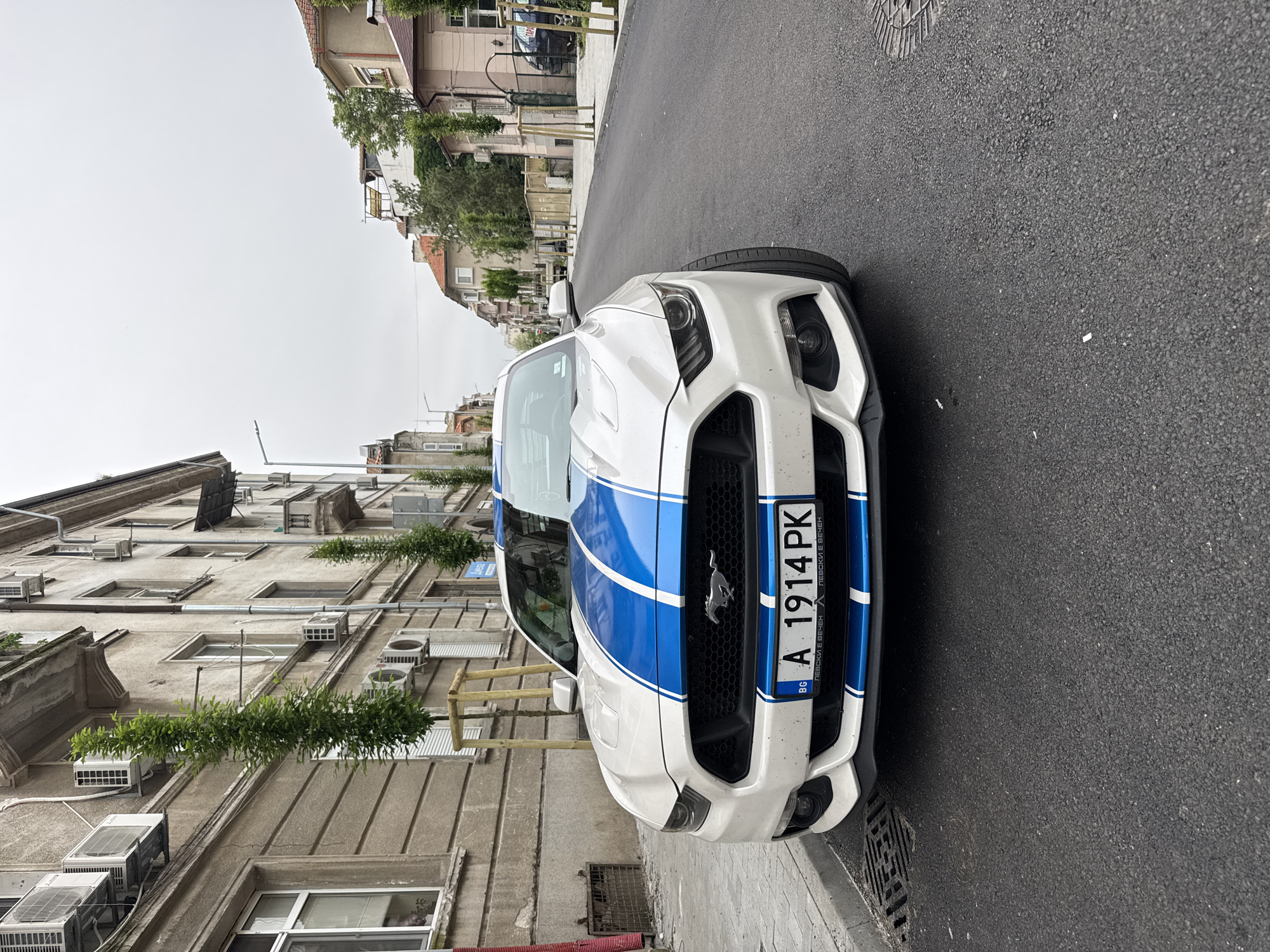The image shows a white Ford Mustang with blue racing stripes parked on the side of a street in Burgas, Bulgaria. The license plate is "A 1914 PK". The street is paved with dark asphalt. On the left is a tall, older building with numerous windows, air conditioning units, and greenery hanging from some of the window areas. Across the street, there are other buildings and trees. The sky is overcast. The car is the main subject, parked on the right side of the frame.