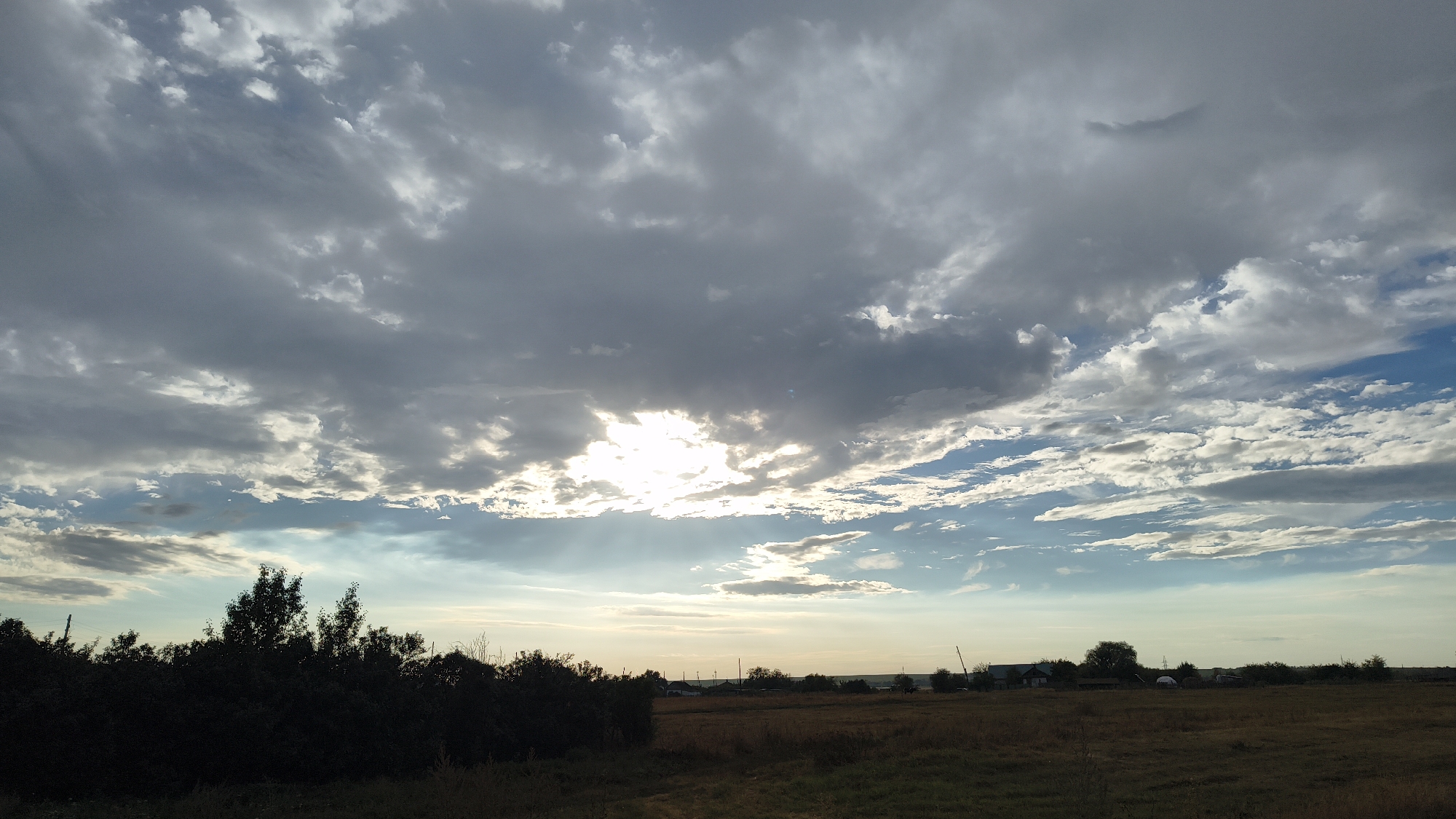 A wide shot captures a rural landscape in Sharashchenskiy, Russia, under a dramatic sky filled with both dark, heavy clouds and patches of bright sunlight. The sun is breaking through the clouds near the center of the frame, casting rays of light downwards. The foreground is dominated by a dark, dense line of trees on the left, with dry, brown grass and a dirt path visible in the lower portion of the image. Beyond the trees, the middle ground reveals a flat expanse of dry, grassy fields with scattered small buildings and trees, suggesting a small settlement. The horizon is low, with the sky occupying the majority of the frame. The lighting suggests it is either early morning or late afternoon.