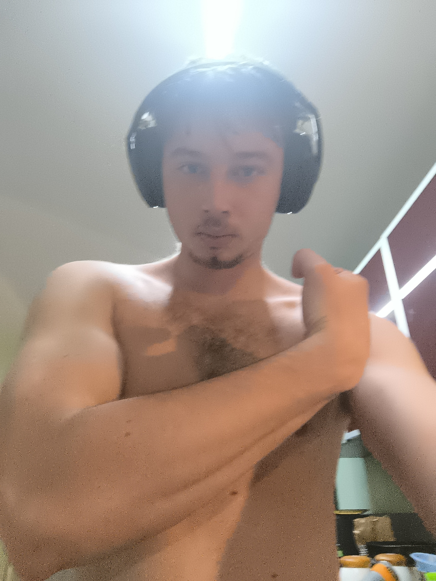 A shirtless man with short dark hair and a goatee is wearing black headphones. He is indoors, likely in a kitchen based on the countertop and objects in the background. A set of cabinets with decorative white trim is visible on the right. The location is unknown and cannot be verified as Sofia, Bulgaria based on the image.