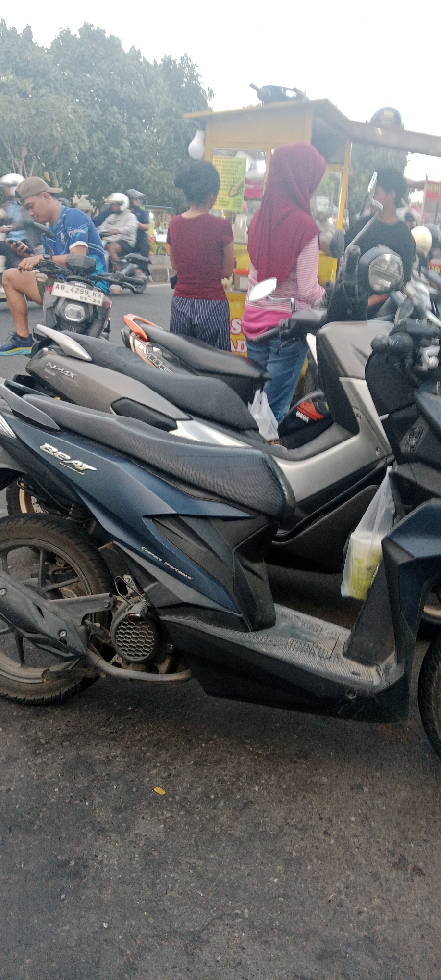 The image depicts a street scene in Ngemplak, Indonesia, with several motorcycles parked or in motion. In the foreground, a dark blue Honda Beat motorcycle is prominently featured, its "BEAT" logo visible on the side panel. To its right, a portion of another motorcycle, possibly a Yamaha NMAX, is visible. Further back, a man wearing a blue t-shirt and a cap is seated on a motorcycle, looking at his phone. Adjacent to him, two women are standing near a yellow food cart. One woman wears a maroon top and striped pants, while the other is wearing a red hijab and a pink and white striped shirt. The food cart has signage with Indonesian text. The environment appears to be a road with some vegetation in the background. The overall scene suggests a typical day in a populated area where motorcycles are a primary mode of transportation.