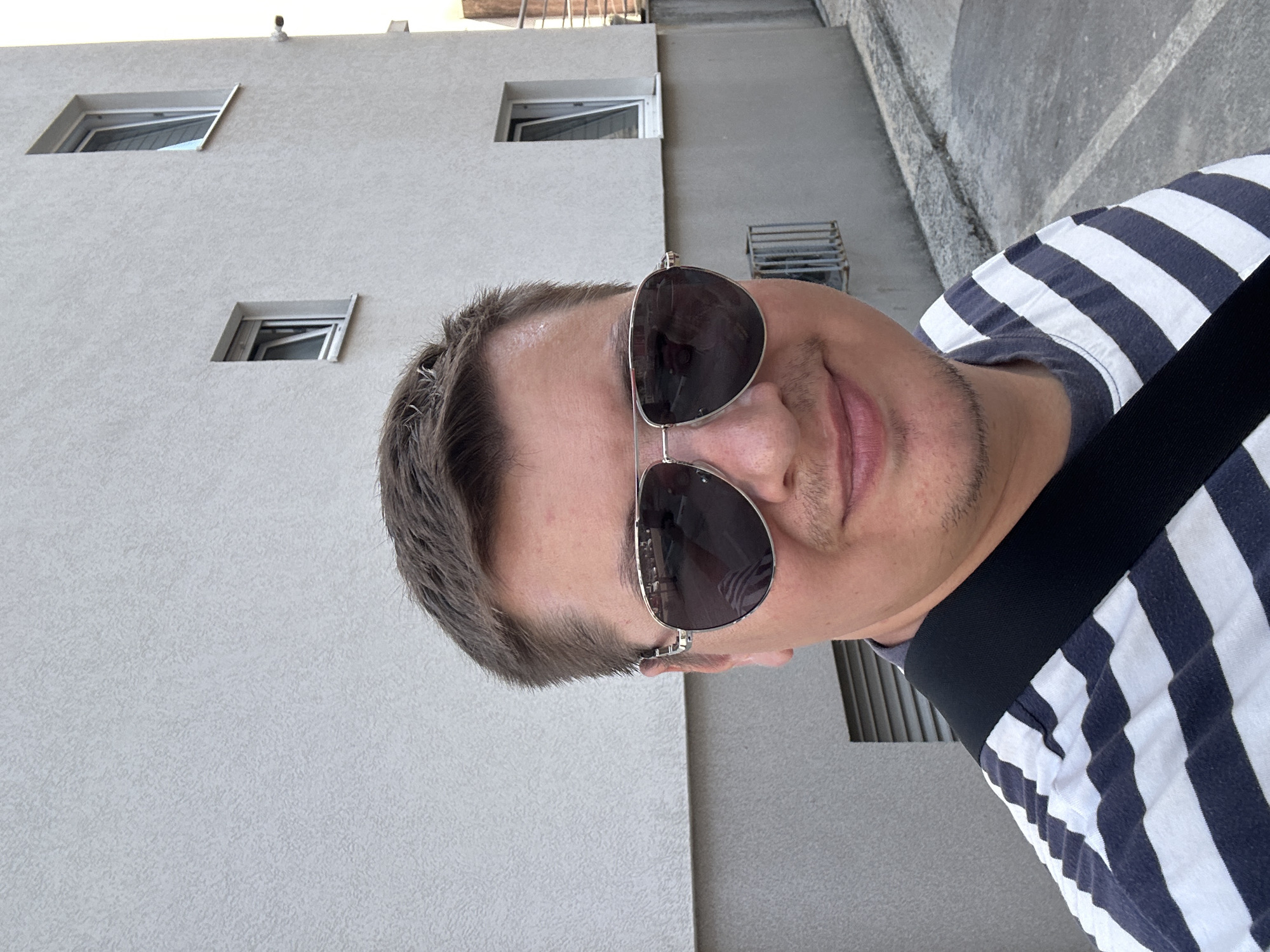 The image shows a person wearing sunglasses and a striped shirt, likely taking a selfie. The person's face is partially obscured by the sunglasses, but they appear to have short, dark hair and light skin. The background consists of a modern building with several small windows and a concrete walkway. The lighting suggests it is daytime, possibly overcast. The overall scene appears to be an outdoor urban or suburban setting. The image is oriented vertically. There is no visible text in the image. The location context provided is Budva, Montenegro.