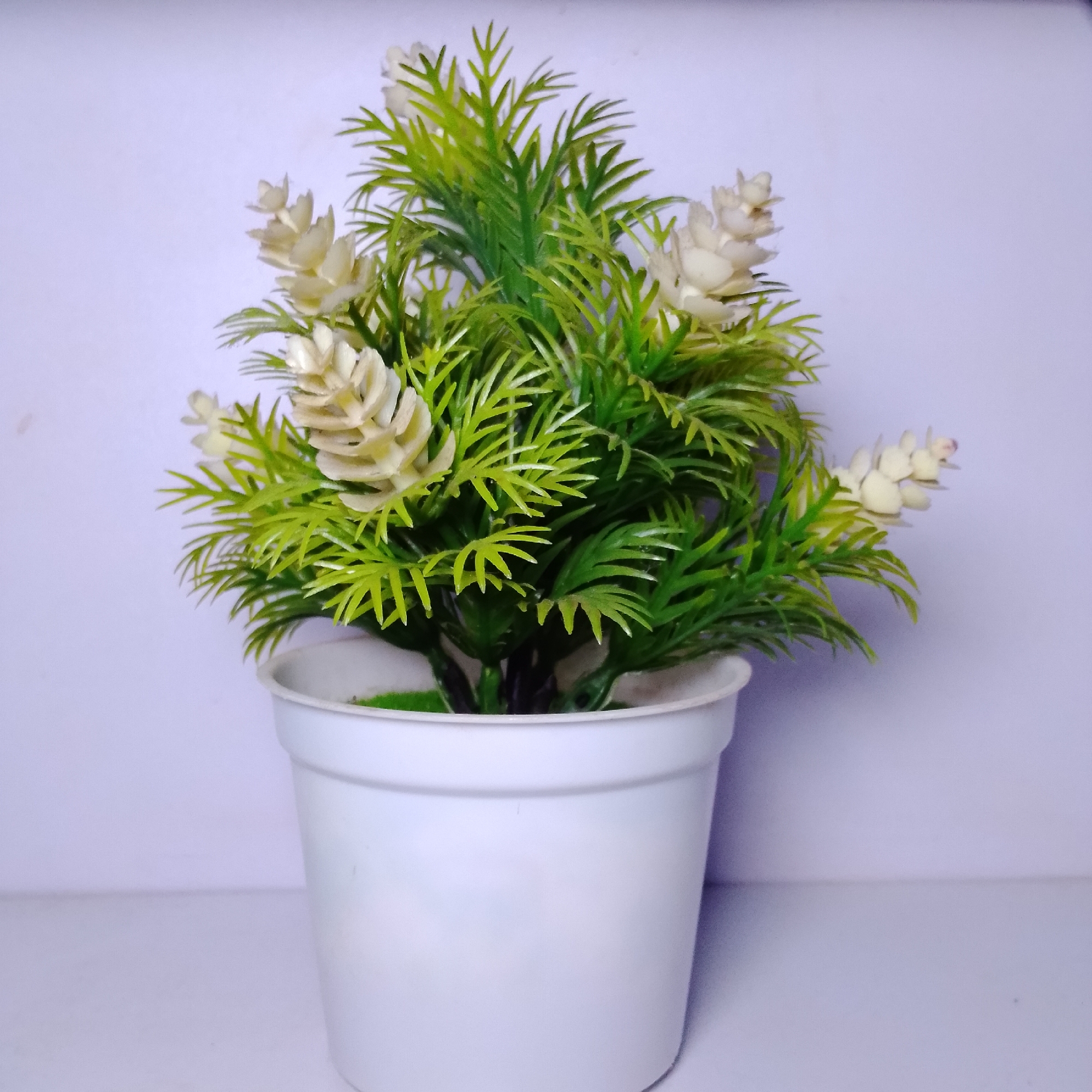 An artificial plant with green, needle-like foliage and small, cream-colored flower spikes is shown in a white plastic pot. The pot is on a white surface, with a light purple background behind it. The image is a close-up, frontal view of the plant.