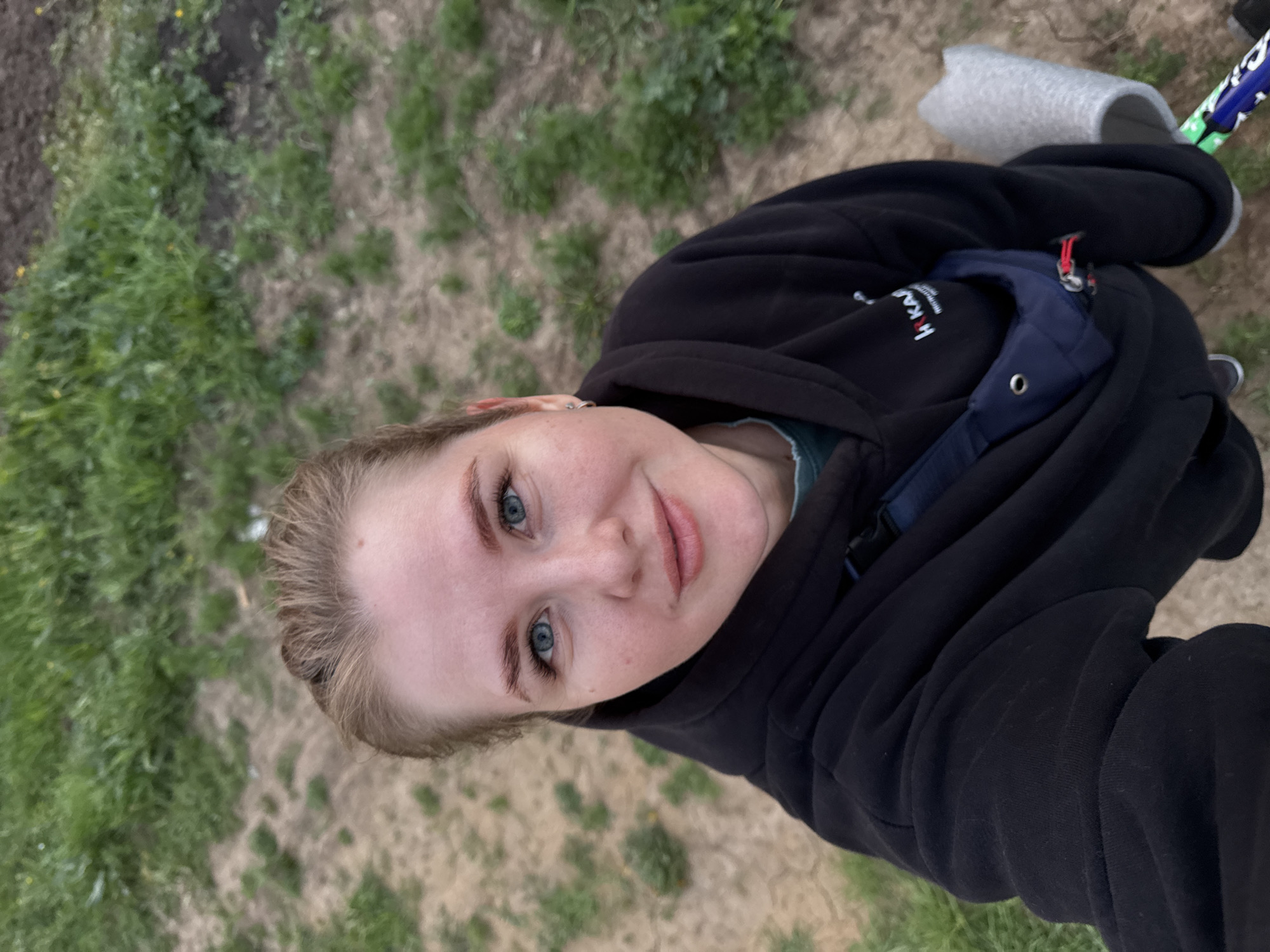 A young woman stands on what appears to be grassy ground, with patches of dirt visible. She is wearing a black hooded sweatshirt and a dark blue fanny pack-style bag across her chest. She has fair skin, light brown hair, and blue eyes. She is looking up into the camera, taking a selfie. Part of a grey slipper-like object and a blue and green object are visible in the upper right corner of the image. The location is likely outdoors, possibly in a park or garden area in Ферма, Russia.