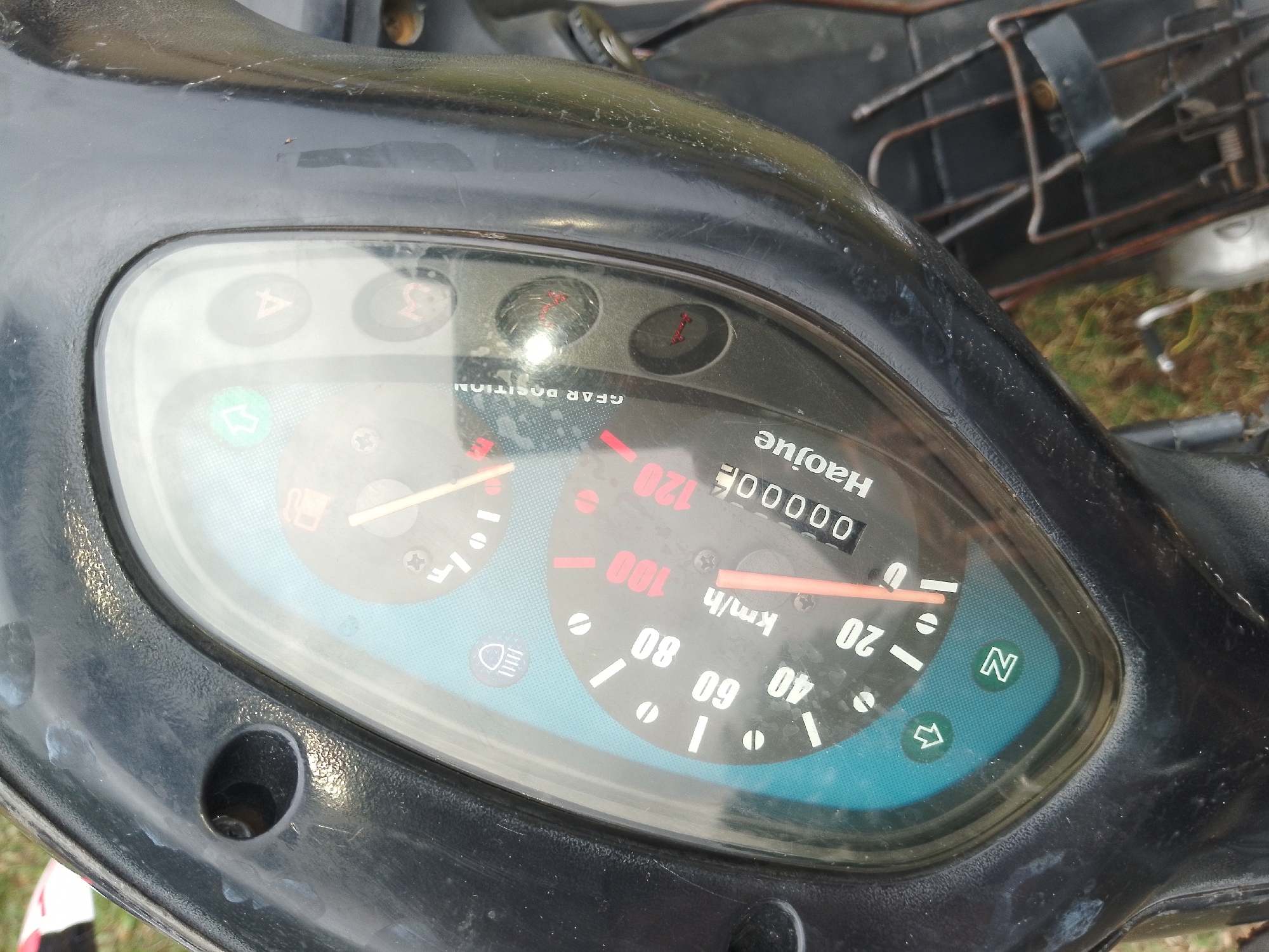 A close-up shot shows a motorcycle's dashboard, featuring a speedometer and other indicators. The speedometer's needle is positioned near the "100" mark, with "KM/H" labeled below. The odometer displays "00000", indicating zero mileage. Several warning lights are visible, including indicators for turn signals, a headlight symbol, and a gear position indicator. A green "N" symbol, likely representing neutral gear, is also present. The dashboard is encased in black plastic, with some scratches and dust visible. The background suggests an outdoor setting with grass and some rustic structures, potentially in Serti, Nigeria.