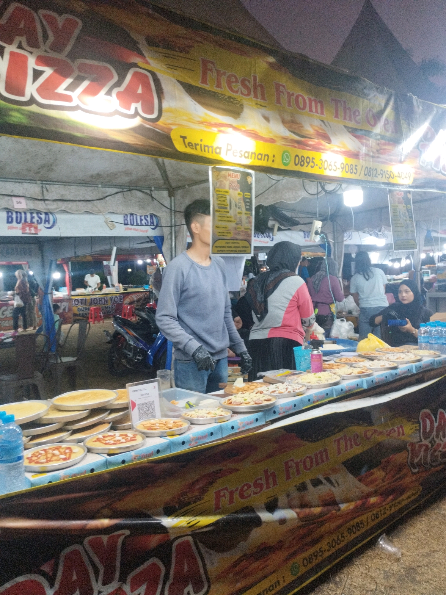 This is a medium shot of an outdoor food stall at night, likely in Parit Padang, Indonesia, selling pizza. A large banner across the front of the stall reads "DAY PIZZA Fresh From The Oven" in large red lettering with "Terima Pesanan: 0895-3065-9085 / 0812-9150-4049" below it. A man in a grey sweatshirt and jeans, wearing black gloves, is standing behind the counter. In front of him is a display of pizzas, some with toppings and some plain, as well as a stack of pizza bases. To the left, a bottle of water is visible. In the background, several people are browsing and ordering food, and a motorcycle is parked. Tents and lighting suggest this is a marketplace or fair.