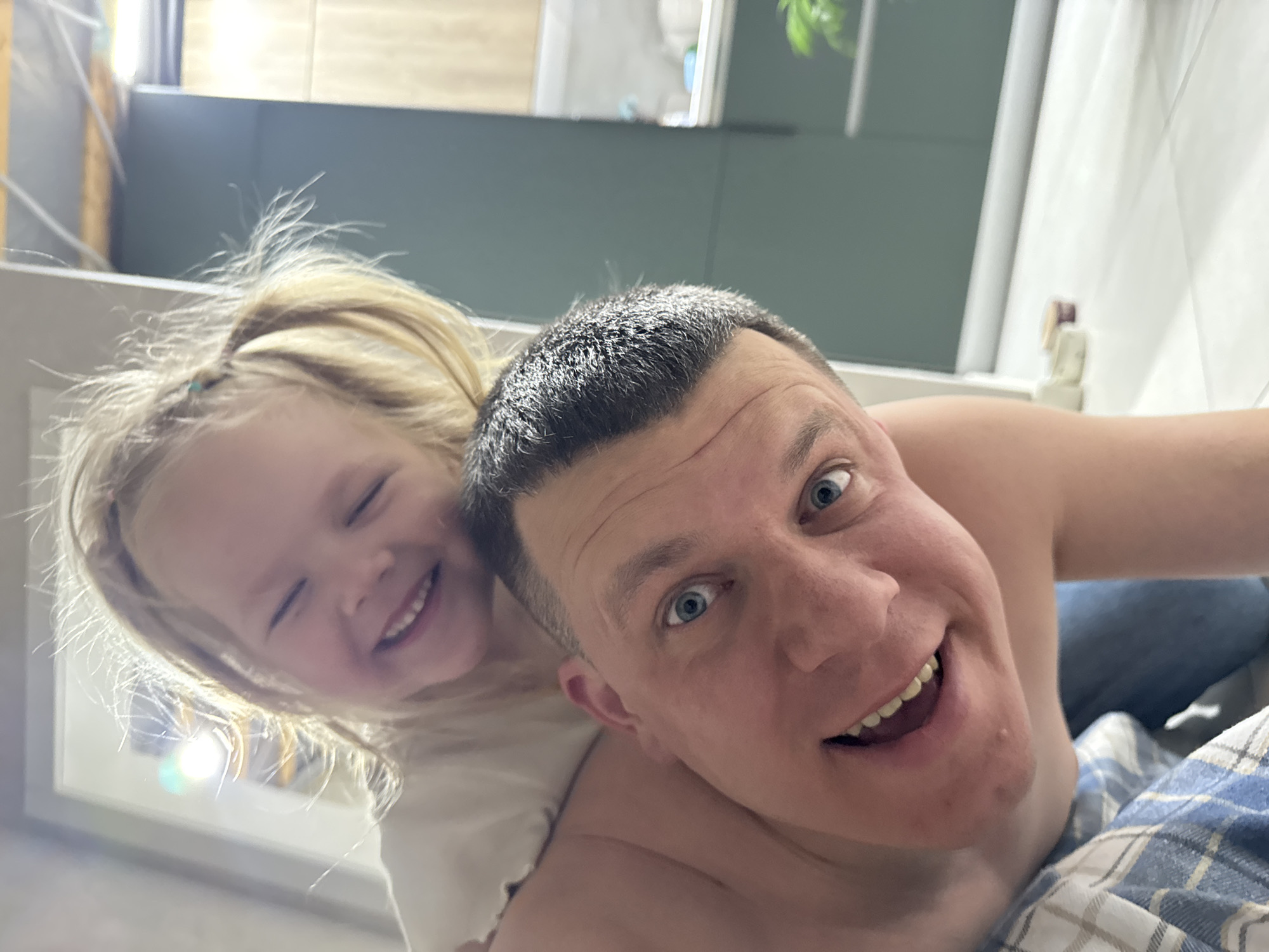 The image shows a selfie taken from a low angle, featuring a man and a young girl. The man has short, dark hair and blue eyes. He is smiling broadly, revealing his teeth. He appears to be shirtless and is wearing blue jeans and a blue and white checked blanket. The young girl, who has light blonde hair pulled back in a ponytail, is behind him and to the left. She is also smiling with her eyes closed slightly. The background includes a mirror, a green wall and a white curtain. The photo appears to have been taken indoors, likely in a bathroom or bedroom. The city of Gornyi (Dvurechenskoe S/P), Russia is not discernible from the image content.