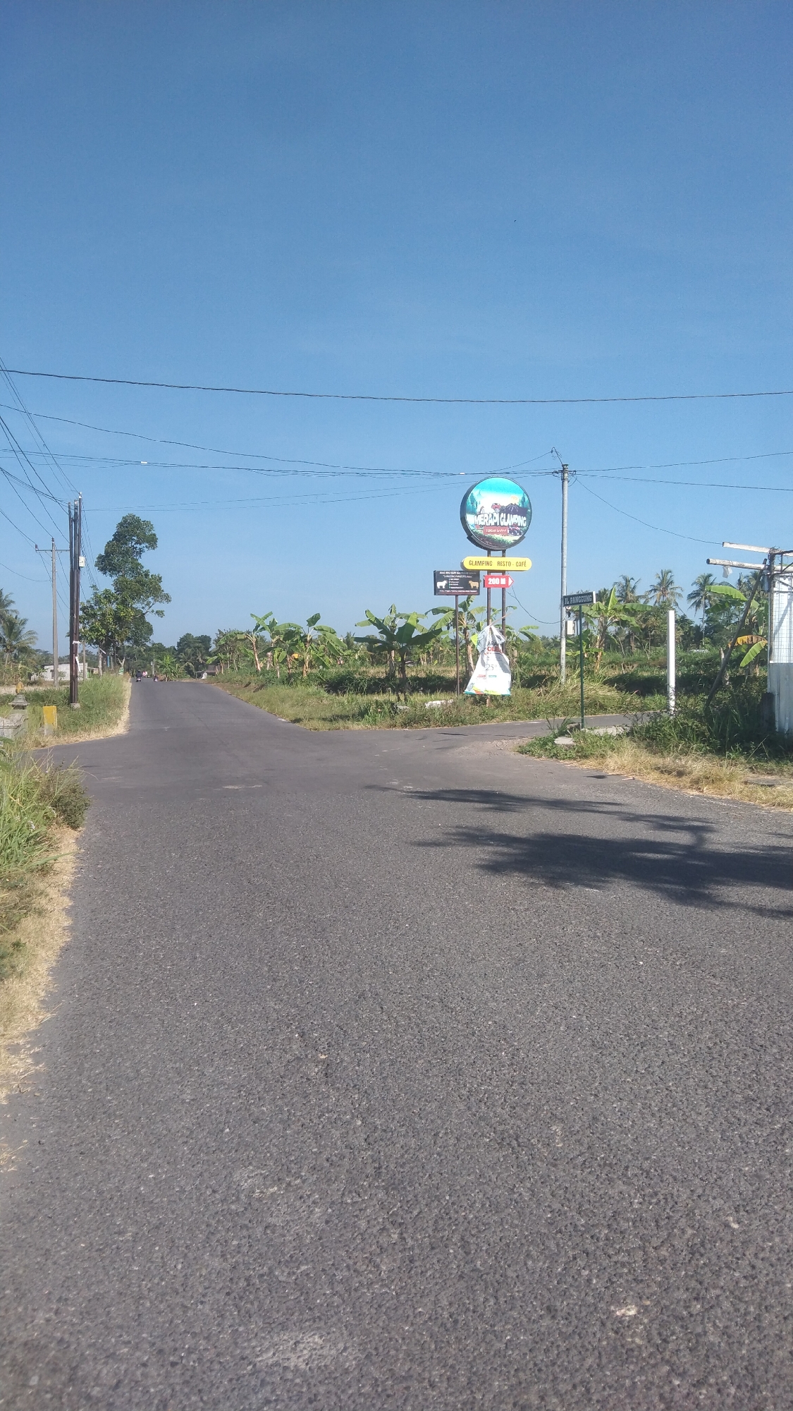 The image depicts a rural road in Argomulyo, Indonesia, under a clear blue sky. The road, paved with asphalt, curves to the left and extends into the distance. To the right of the road, there is a sign that reads "GLAMPING RESTO CAFE" with an arrow pointing towards it and the distance "200 M". Above this sign is a larger circular sign with "MERAPI CLAMPING" written on it, featuring a scenic landscape. A white plastic bag with colorful markings hangs from a pole near the signs. The surrounding environment is characterized by abundant vegetation, including banana trees and palm trees, indicative of a tropical or subtropical climate. Power lines and poles are visible on the left side of the road. The scene is devoid of people.