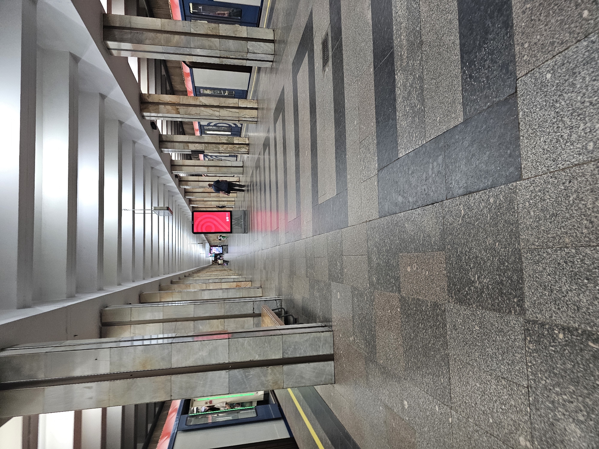 This is a low-angle, wide shot of a metro station platform in Moscow, Russia, likely on the Filyovskaya Line due to the train design. The station features a long, covered platform with a series of massive, square marble-clad pillars supporting a white, barreled ceiling. The floor is paved with granite tiles, some of which are dark grey and arranged in a geometric pattern.
On the left, a train with blue and white carriages is stopped at the platform, its doors open. A yellow line marks the edge of the platform. On the right side of the platform, more train carriages are visible, also stopped.
A single man, wearing a dark shirt and pants, walks away from the camera towards the far end of the platform. Further down the platform, several other indistinct figures are visible, suggesting other passengers. A large, bright red digital advertisement is mounted on a pillar in the middle distance, displaying a swirling pink and white logo. A wooden bench is positioned between two pillars near the foreground. The overall atmosphere is utilitarian and spacious, typical of Soviet-era metro architecture.
