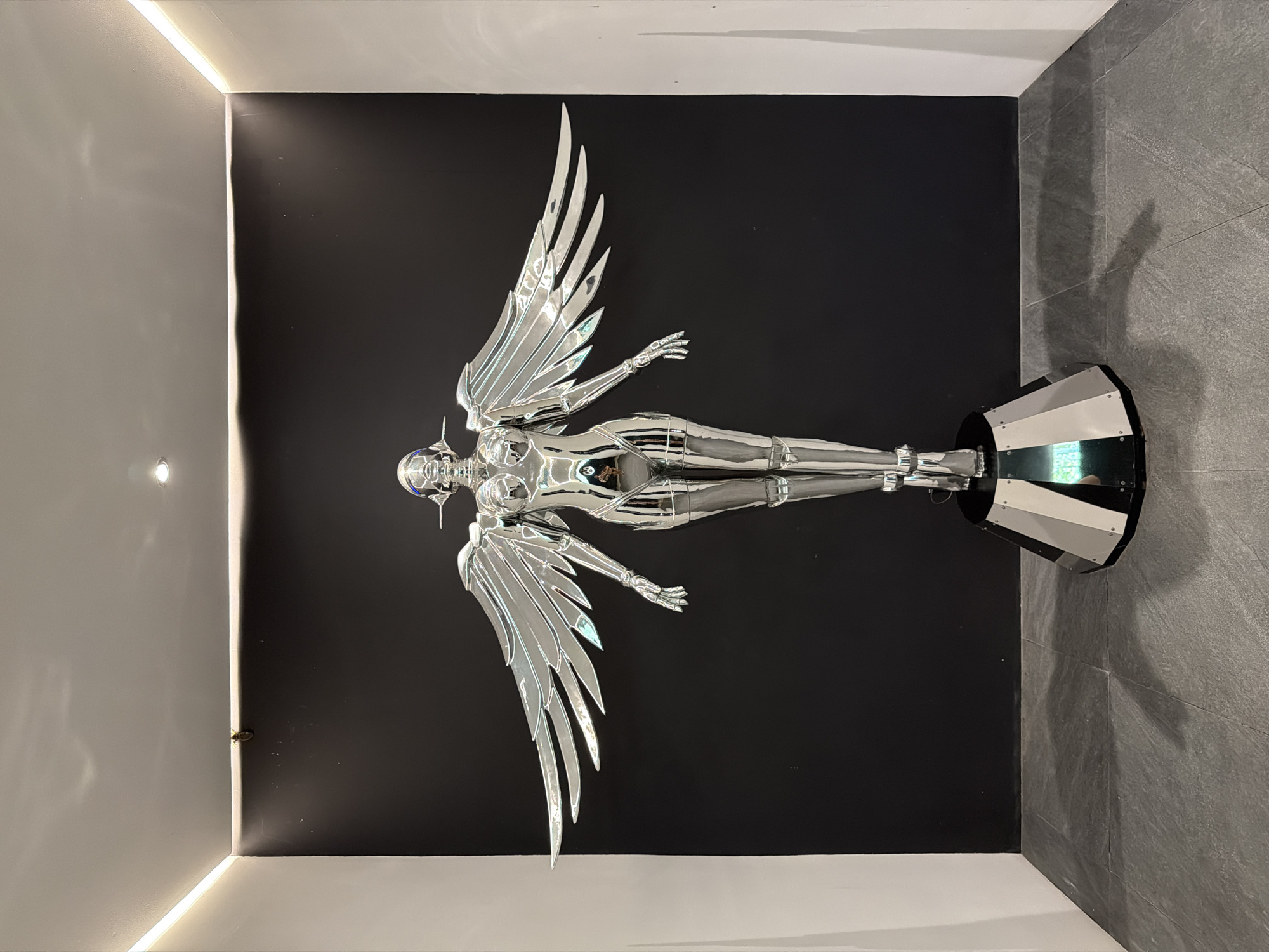 The image shows a full-body shot of a metallic, angel-like sculpture with large, spread wings, positioned in the center of a room. The sculpture has a humanoid form and is highly reflective, casting a bright sheen. It stands on a multi-faceted, metallic base. The background consists of a plain black wall with white borders on the left and right, and a white ceiling with recessed lighting. The floor is tiled with grey, stone-like material. There is a reflection visible on the sculpture's abdomen, appearing to be a person or another object. The scene is well-lit, with the lighting highlighting the metallic surface of the sculpture. This artwork is located in the city of Ko Samui District, Thailand.