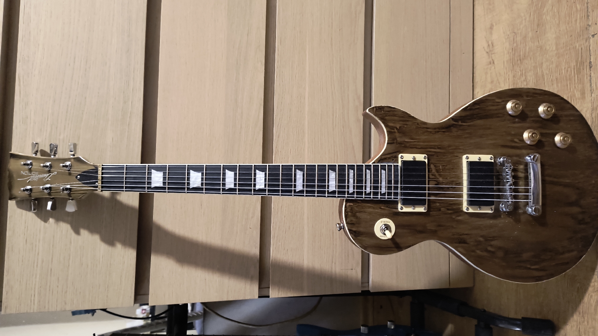 A Les Paul style electric guitar rests horizontally on a light-colored wooden surface with vertical paneling. The guitar's body has a wood grain finish, with cream-colored binding around the edges. It features two humbucker pickups with cream-colored pickup rings, and gold-colored control knobs. The headstock also has a wood finish, with tuning pegs and a signature. The guitar's fretboard is black, with white trapezoidal inlays. Below the wooden surface, dark cables, and a black tripod stand are visible. The location is unknown and there is no indication in the image that it is Sofia, Bulgaria.