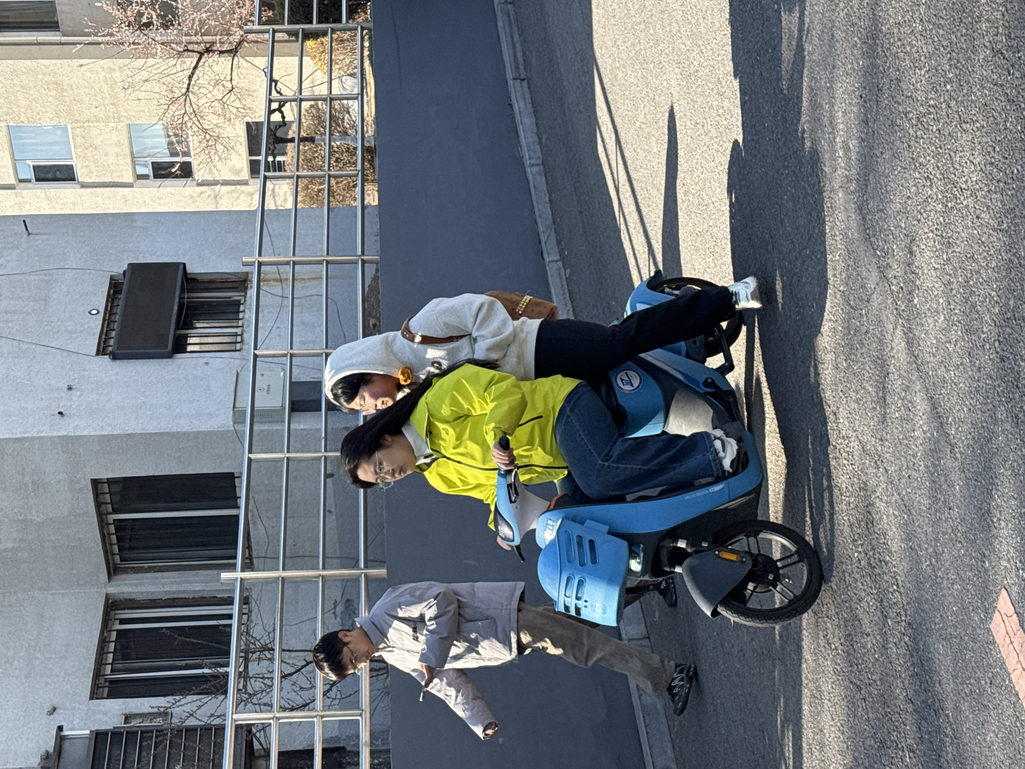 Two people are riding a blue electric scooter on a paved road in Zhongtun, China. The scooter is identified by the "HELLO DESIGN" insignia on the side. A third person is walking beside the scooter. The scooter driver wears a bright yellow jacket and jeans. The passenger is wearing a light-colored hoodie and black pants, with a brown purse over her shoulder. The person walking wears a light-gray jacket and brown pants. A building with windows and a metal fence is in the background. The weather appears sunny.