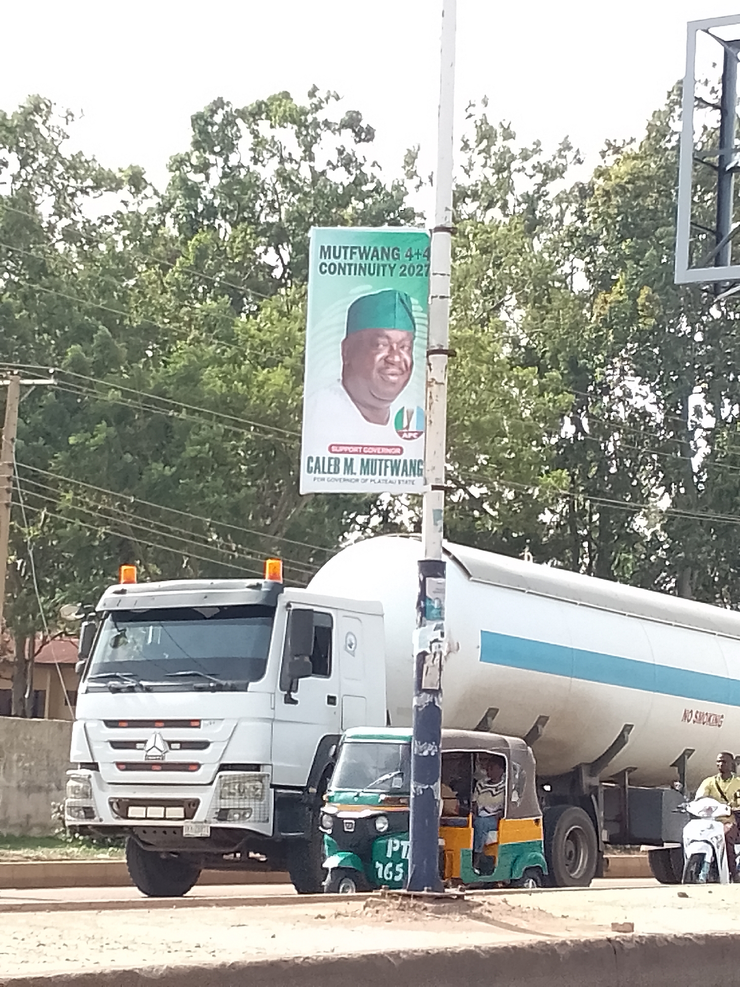 The image shows a street scene in Bukuru, Nigeria. A political banner promoting Caleb M. Mutfwang for governor of Plateau State hangs from a utility pole. The banner features Mutfwang's image and states "MUTFWANG 4+4 CONTINUITY 2027". In the foreground, a white tanker truck dominates the view, obscuring much of the background. A green and yellow auto-rickshaw (tuk-tuk) is parked near the truck. A man is driving a scooter next to the tanker. Trees are visible in the background.
