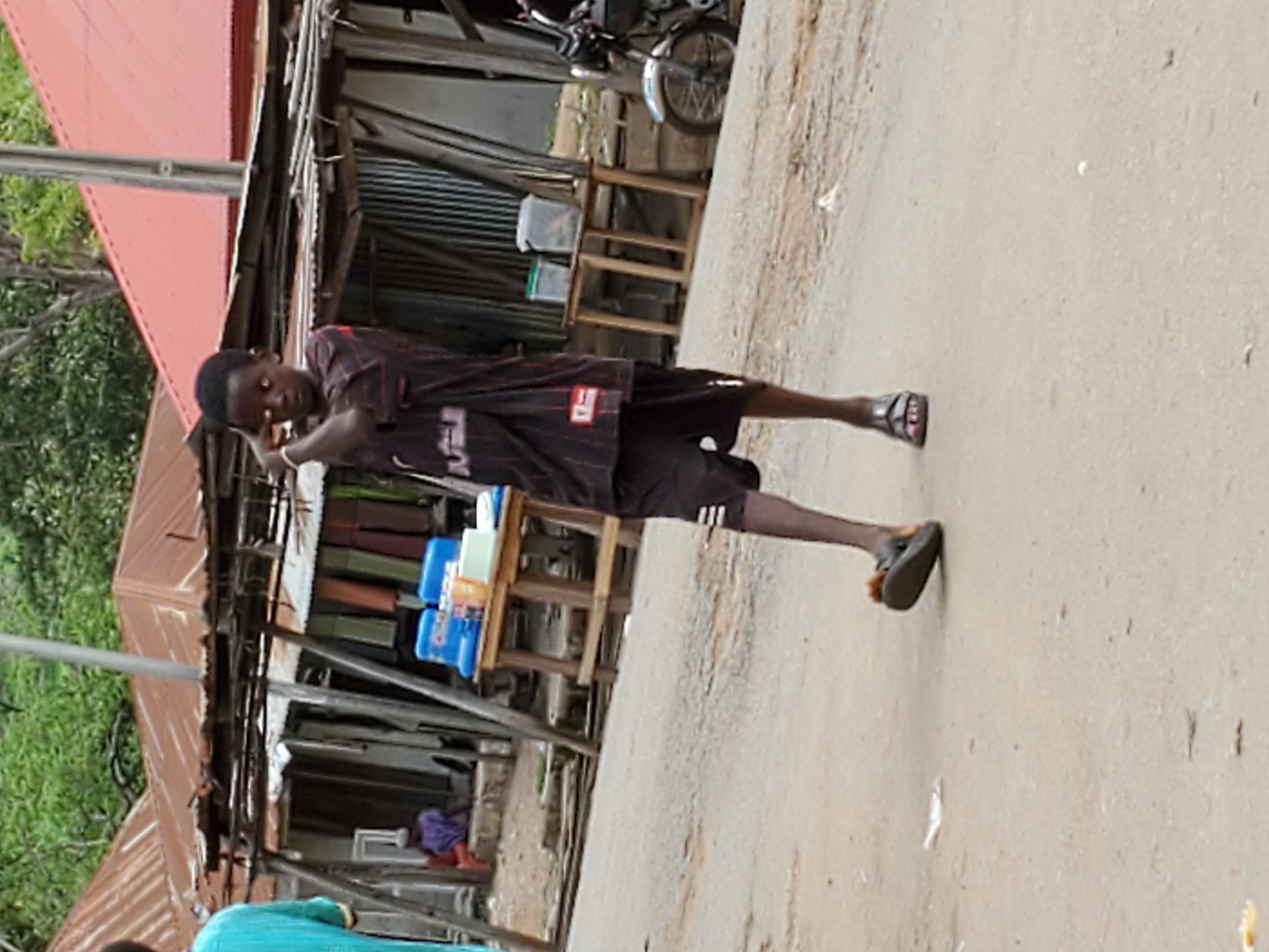 The image depicts a person of African descent, appearing young, standing on a paved or dirt road. They are wearing a dark, short-sleeved shirt with red and white accents, dark shorts, and sandals. The individual is striking a pose with their hands near their head.
Behind the person is a building constructed of corrugated metal with a rusted or reddish roof. The building appears to be a small shop or stall, with various items displayed on makeshift shelves. These items include packaged goods, possibly cleaning supplies, in blue containers.
Further in the background, there is vegetation and a motorcycle parked next to the building.
The scene is set in Mararaba, Nigeria, suggested by the style of the building and general appearance of the environment.