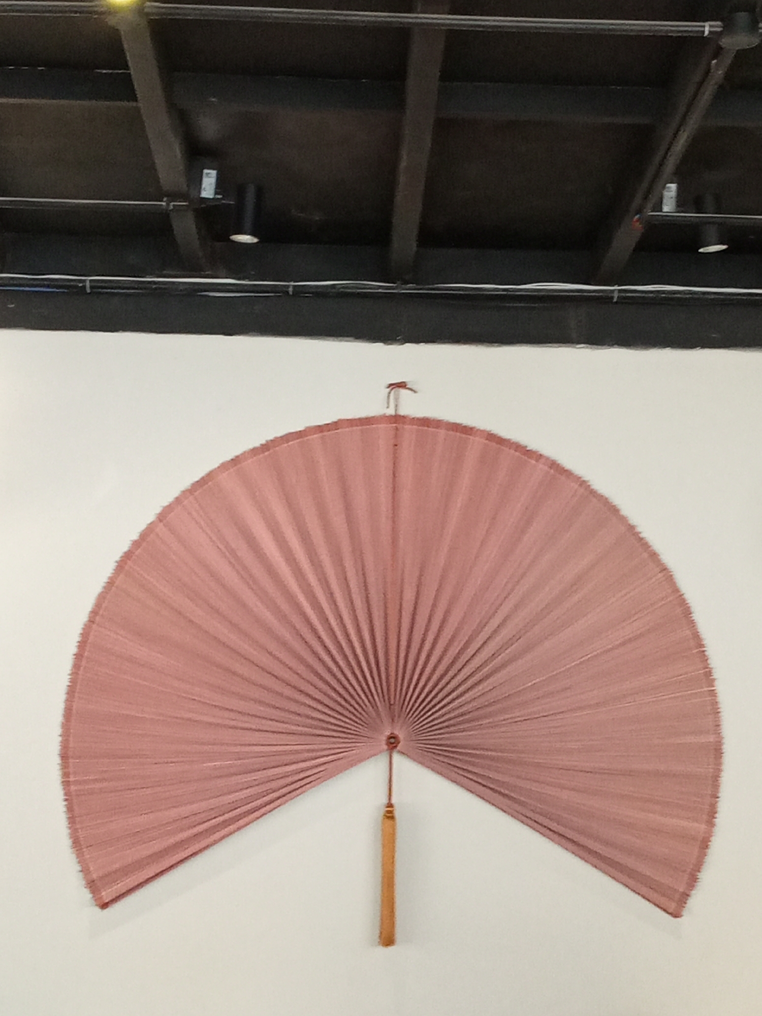 A large, semi-circular decorative fan, composed of numerous radiating pleats in a dusty rose or mauve hue, is centrally mounted on a plain white wall within an interior space in Nha Trang, Vietnam. The fan's woven material, possibly natural fiber, displays an irregular textured edge along its circumference. A small, reddish-brown wire or hook is visible at the top center, securing the fan. From the central bottom pivot point of the fan, a slender, rectangular golden-brown tassel hangs vertically. Above the white wall, an exposed ceiling features dark structural beams and joists, with multiple cylindrical black light fixtures and white electrical conduits visibly mounted across the dark surface. No people are present in the scene.