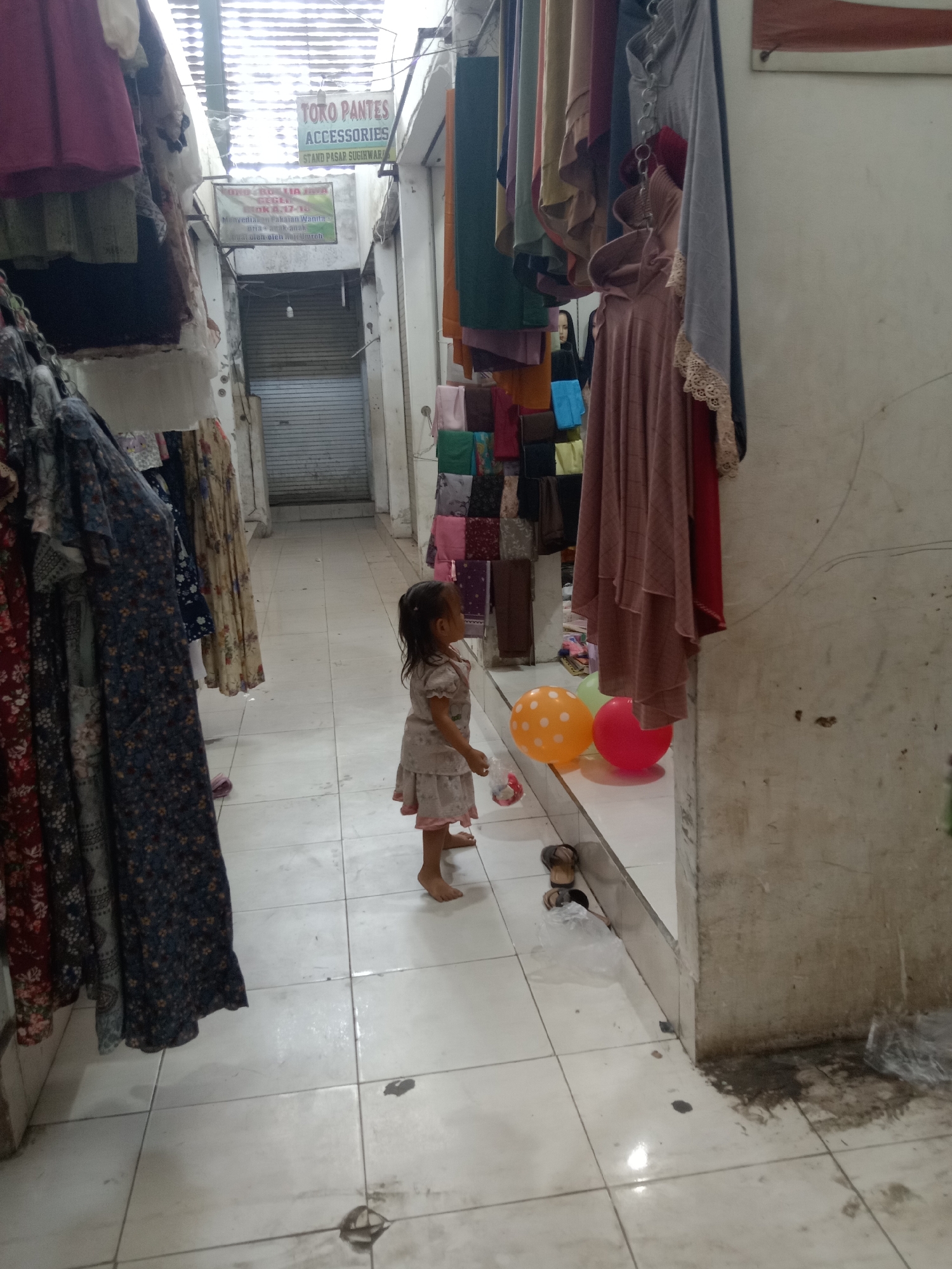 The image depicts a narrow indoor market aisle in Sugihwaras, Indonesia, with clothing stalls lining both sides. A young girl, wearing a light-colored dress, stands in the center of the aisle, looking to her right. In front of her are three balloons: one orange with white polka dots, one light green, and one red. To the right of the balloons and slightly behind them, a small, dark object resembling a toy or a doll is visible. The aisle floor is tiled and appears slightly wet in places. Hanging clothes, primarily dresses and hijabs, are displayed on racks. A sign above the stalls reads "TORO PANTES ACCESSORIES STAND PASAR SUGIHWART." The lighting is dim, suggesting an indoor, possibly covered, market environment.