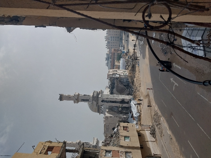 The image displays a scene of widespread urban destruction in Khan Yunis, Palestinian Territory, captured from an elevated perspective. In the midground, a large mosque with a prominent minaret and a partially collapsed dome stands amidst significant structural damage. Surrounding the mosque are numerous multi-story buildings, extensively damaged or reduced to piles of rubble, exposing structural rebar and debris.
A paved road with visible lane markings (including arrows for turns) extends through the foreground and midground. The ground adjacent to the road and surrounding the damaged buildings is covered in debris, dust, and sand. Several individuals are visible across the scene: some walk on the rubble-strewn areas near the destroyed buildings, others are seen further down the road, and one person stands alone on the debris near the road's center. In the mid-right, a donkey-drawn cart is positioned on the roadside, attended by multiple people. The sky is hazy or overcast. A dark, ornate metal railing or balcony fixture occupies the immediate foreground, partially obscuring the lower right of the frame.