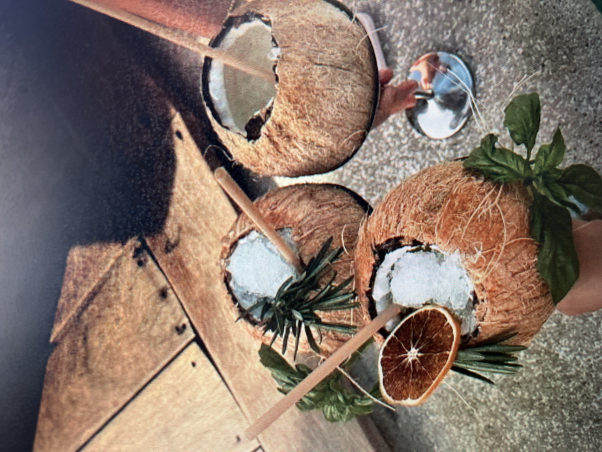 The image shows three coconuts filled with a white beverage and garnished. One coconut is in a stemmed glass, suggesting it is a drink. Another coconut is placed on a textured surface, possibly a wooden deck or floor, and garnished with a sprig of rosemary and a slice of dehydrated orange. A third coconut, also on the textured surface, is garnished with green leaves, possibly mint or basil. Wooden planks with visible nails and dark shadows are visible in the background. The overall setting appears to be outdoors, with natural lighting casting shadows. The image does not provide any specific information or visual cues to pinpoint a location within Sejahtera, Indonesia.