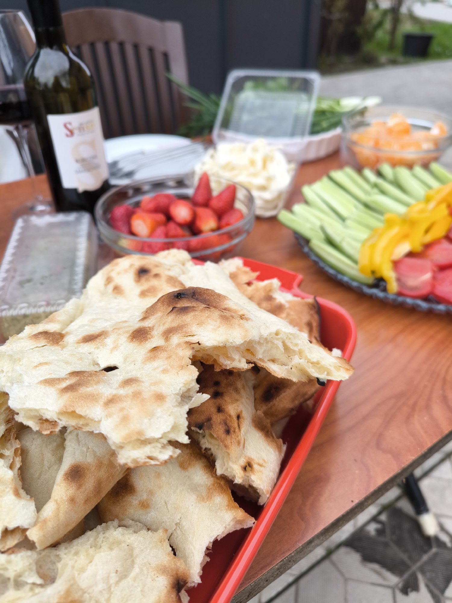 The image presents an outdoor dining setup. A red bottle of wine stands beside a glass, along with a white plate and cutlery. A bowl of strawberries, a bowl of what appears to be whipped cream, a container of greens, a bowl of sliced oranges, and a platter of celery sticks, yellow peppers, and sliced tomatoes are arranged nearby. In the foreground, a red dish overflows with flatbread pieces. The scene takes place on a wooden table, likely in a garden or patio setting. The background features a grey wall and glimpses of greenery, suggesting an outdoor location. The environment is consistent with a gathering of people for food and drinks. The picture was taken in Novofryazino, Russia.