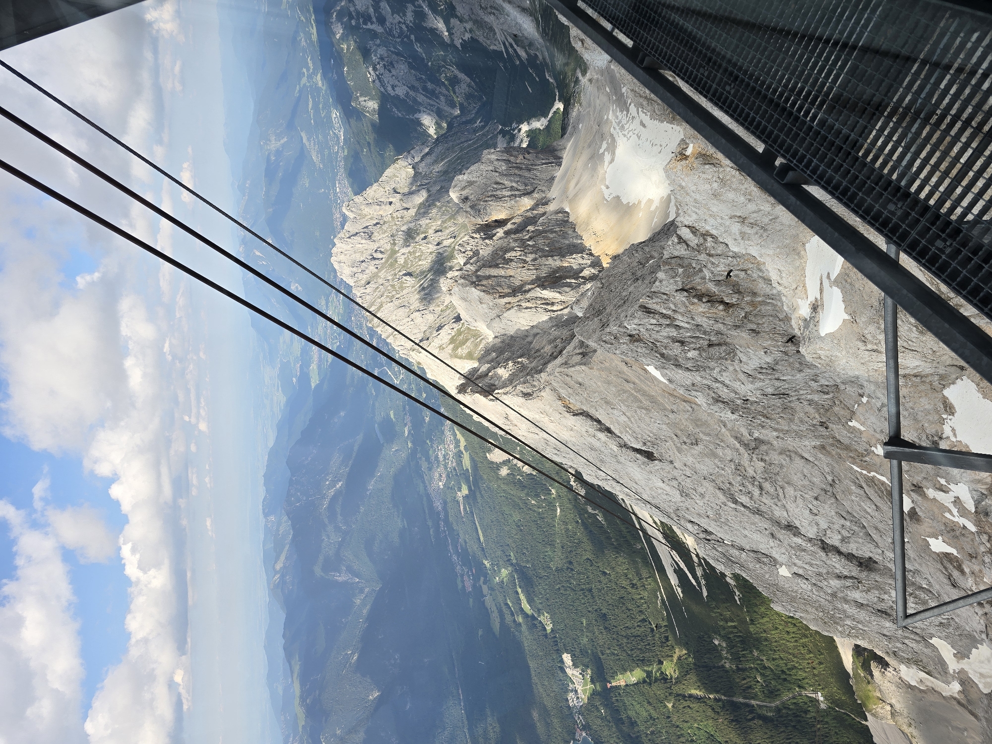 The image captures a high-altitude view of the Zugspitze mountain range near Garmisch-Partenkirchen, Germany, as seen from a cable car. Several thick cables, part of the cable car system, stretch diagonally across the frame, disappearing into the upper right corner. Below, the rugged, rocky terrain of the mountains is dominant, with sharp peaks and deep valleys. Patches of snow are visible on the shaded slopes.
In the distance, a sprawling valley unfolds, dotted with evergreen forests. A town or village can be seen nestled in the valley, with buildings and roads. The landscape extends to a hazy horizon of rolling mountains under a bright blue sky with scattered white clouds. A metal grated walkway or platform, likely part of the cable car station or a viewing area, occupies the lower right portion of the image, providing a man-made element to the natural grandeur. No people are visible in the frame.