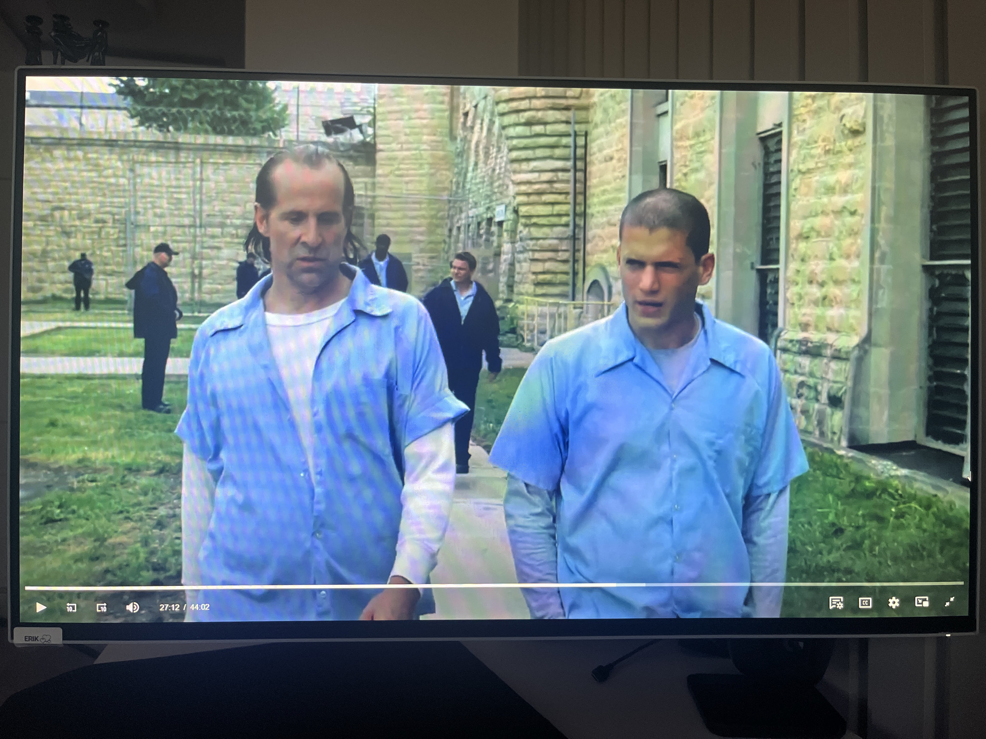 The image displays a television screen showing a scene from a prison. Two male subjects, appearing to be inmates, are walking towards the viewer. They are both wearing light blue button-down shirts over white undershirts, typical prison attire. The subject on the left has a receding hairline and is looking forward with a neutral expression. The subject on the right has a shaved head and is also looking forward, with a slight furrow in his brow. In the background, other figures in dark clothing, possibly guards or other inmates, are visible. The environment appears to be an outdoor prison yard with a stone building and a chain-link fence. The bottom of the screen shows video playback controls with timestamps and icons. The television itself is mounted on a stand, and the image quality is typical of a video playback on a screen. There is no indication that this scene is located in the city of Varna, Bulgaria.