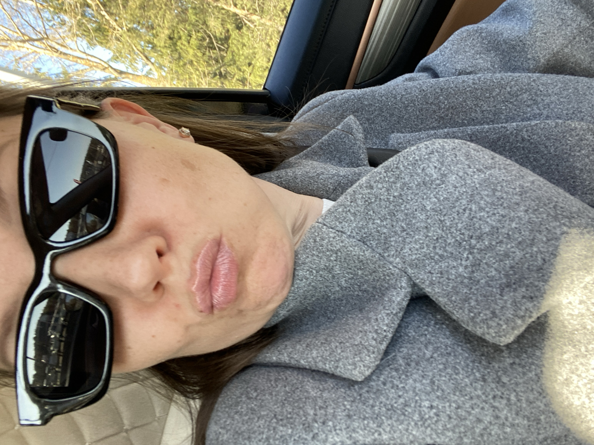 A woman wearing sunglasses and a gray coat is in a car. Outside the car window, trees are visible. The location in the city of Chaikovsky, Russia is not discernible from the image.