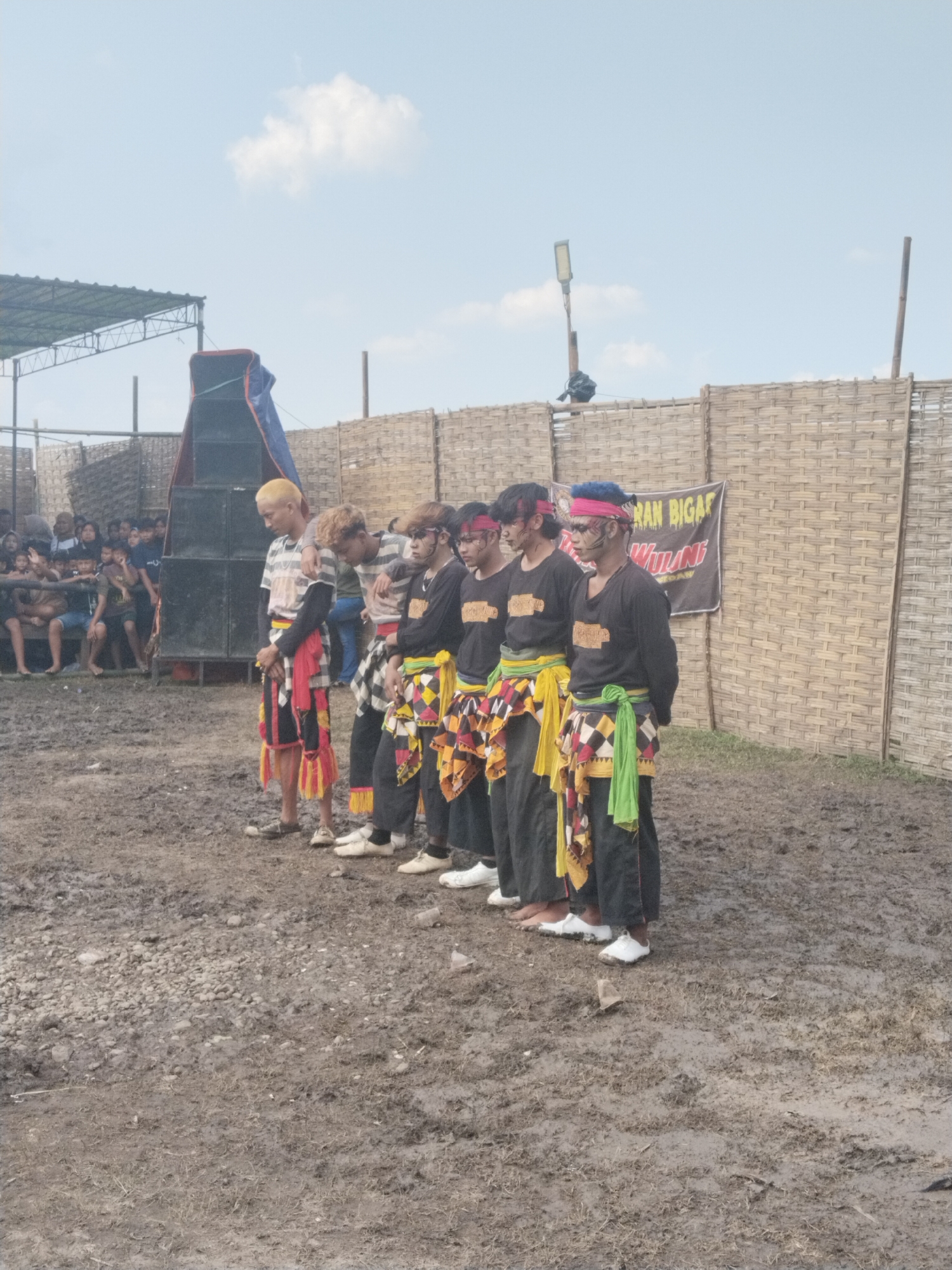 This image captures an outdoor cultural performance in Kedawung, Indonesia, under a bright, partly cloudy daytime sky. In the foreground, a group of approximately seven young men stand in a line on muddy and gravelly ground. They are dressed in black long-sleeved shirts, black trousers, and colorful patterned sarongs or wraps around their waists, featuring shades of yellow, red, green, and black and white checks. Several performers have headbands and distinctive face paint, indicating their roles in the event.
To the left, large black speakers, partially covered by a blue tarp, are set up next to a makeshift canopy structure where an audience is gathered, observing the proceedings. The background features a long fence made of woven bamboo, which stretches across the scene, providing a rustic backdrop. A light fixture on a pole is visible above the fence.
A banner attached to the bamboo fence displays the text "JARAN BIGAR WULUNG." This suggests the event is a traditional Javanese horse dance performance, possibly related to Jaranan or Kuda Lumping, known for its energetic nature ("Bigar" implies lively) and often featuring black (Wulung) elements. The muddy and uneven ground signifies an unpaved, open-air setting, common for community cultural events. The performers appear focused, poised for or participating in the traditional dance.