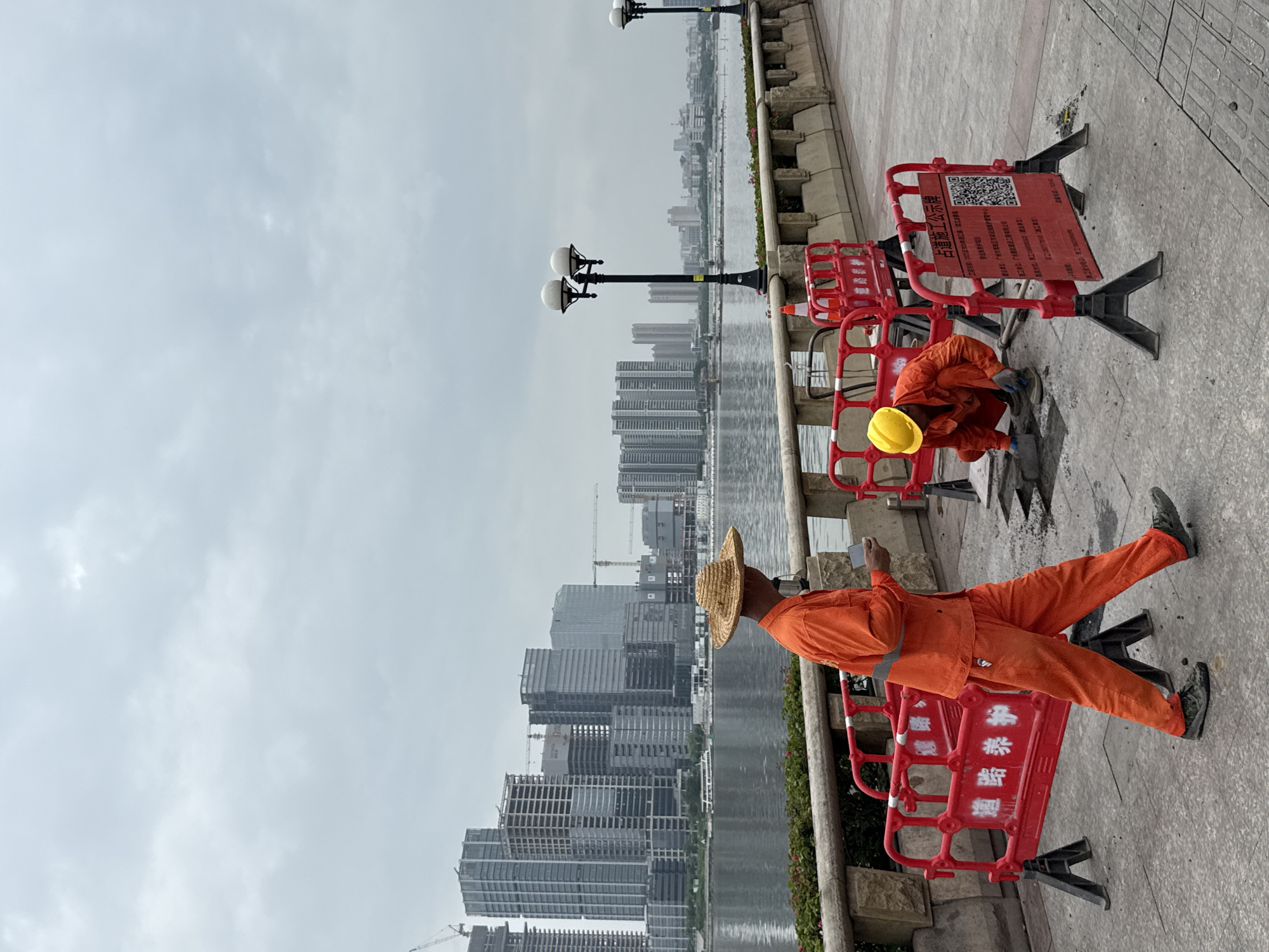 The image depicts an overcast day scene along a waterfront promenade in Haizhu District, China, with a cityscape of modern buildings in the background. Two construction workers are present. One worker, wearing an orange uniform and a straw hat, is walking away from the camera, looking at a mobile phone in his hand. The second worker, in an orange uniform and a yellow hard hat, is kneeling and appears to be working on the paved ground, possibly repairing or installing something. Red construction barriers with Chinese text are placed around the work area. The promenade is paved with stone tiles. Beyond the promenade, a body of water separates the foreground from the backdrop of numerous high-rise buildings, some of which are under construction, indicated by cranes. Streetlights are visible along the promenade.