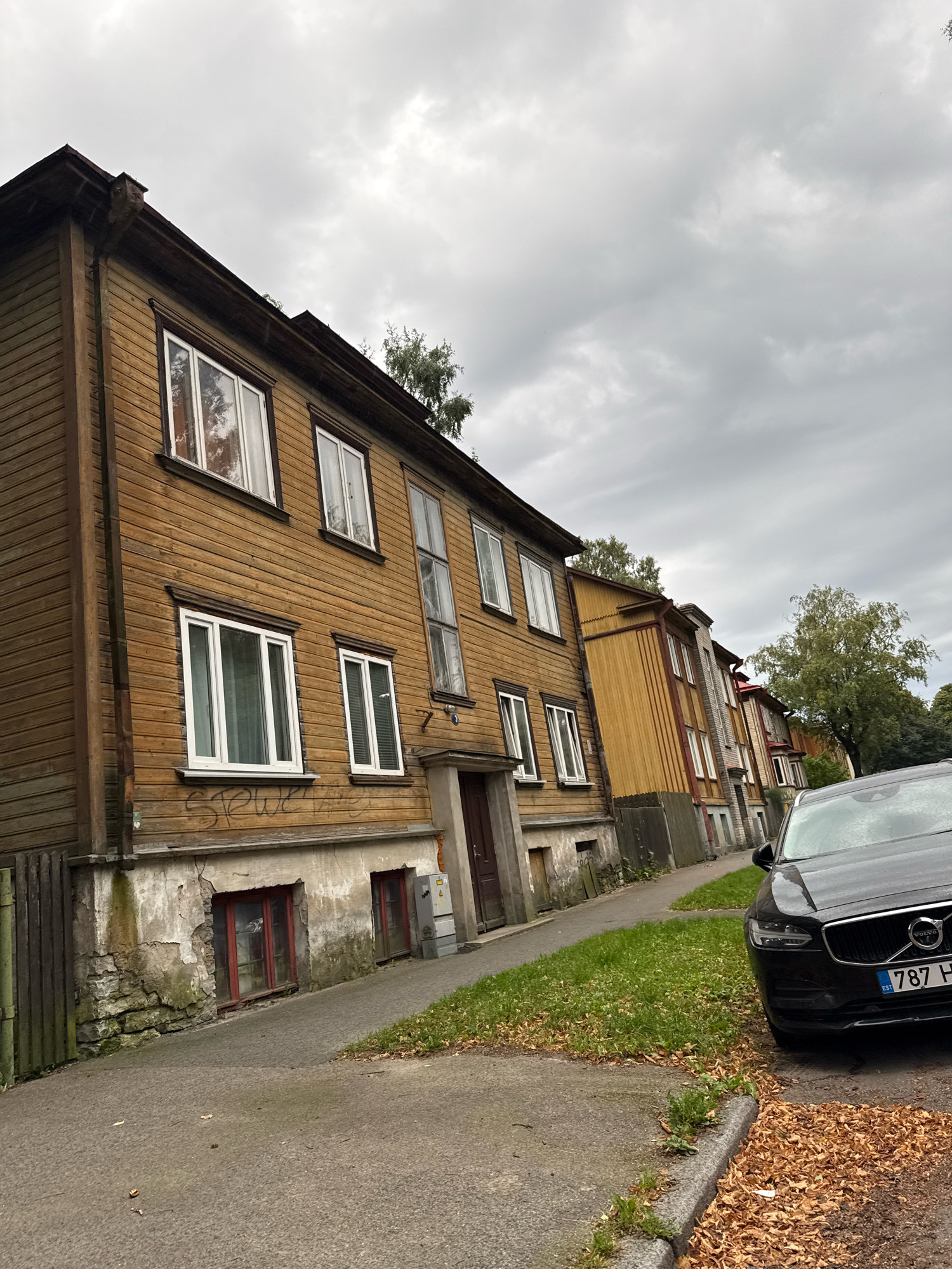 The image captures a street scene in Tallinn, Estonia, featuring a row of weathered wooden residential buildings lining a sidewalk and a paved road. The buildings, primarily of a brown and yellow hue, display traditional architecture with visible wooden siding and numerous windows. The sky above is overcast, contributing to a muted, cloudy atmosphere. A dark-colored Volvo SUV is parked on the right side of the road, facing away from the camera, with its license plate visible. Fallen autumn leaves are scattered on the ground around the parked car and along the edge of the sidewalk. The buildings show signs of age, with some visible graffiti on the lower section of the leftmost structure and some wear and tear on the facade of the buildings. The scene appears to be a quiet residential area.