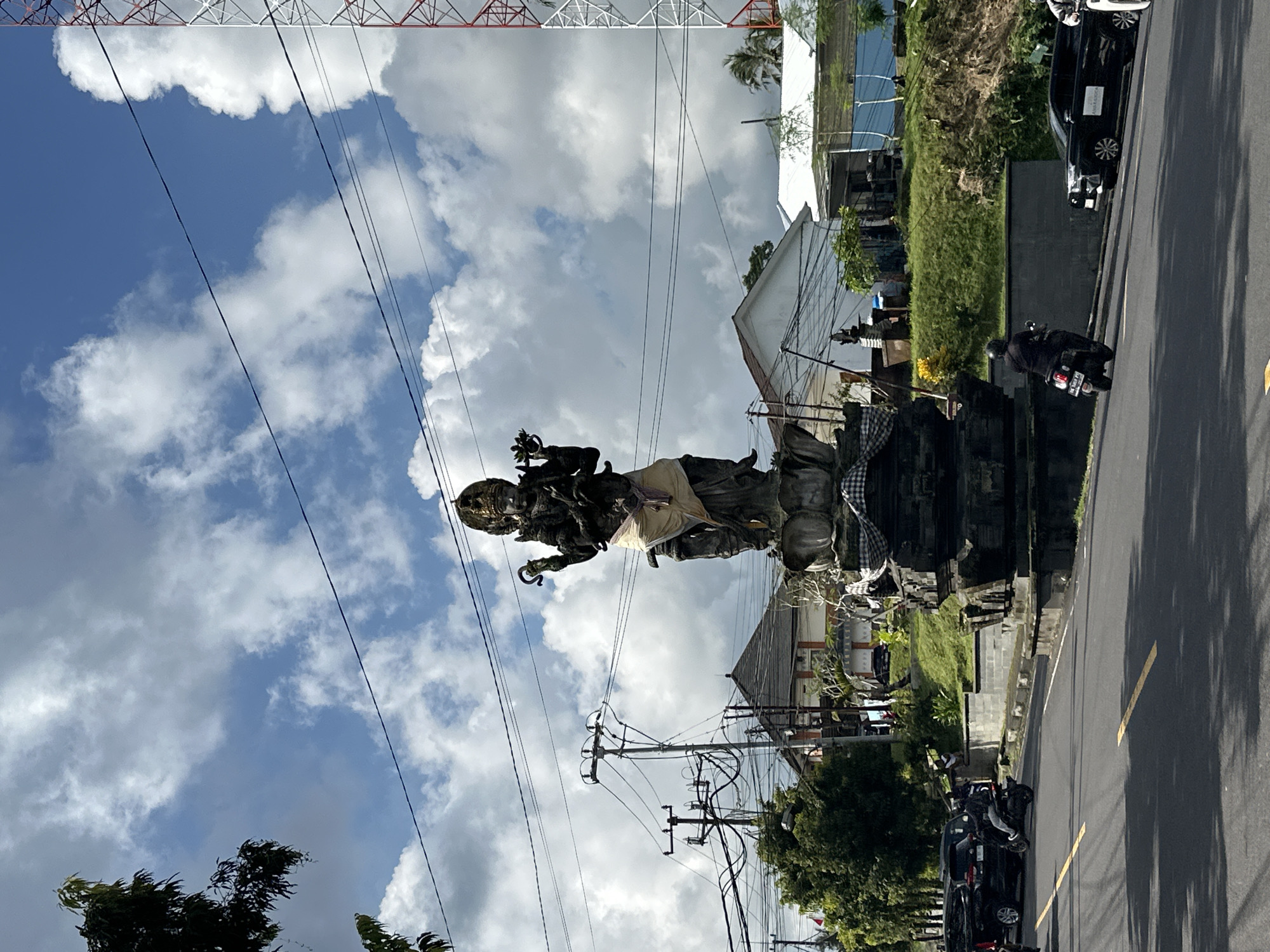 The image shows a street scene in Tampaksiring, Indonesia. A statue of a deity stands on a pedestal adorned with a black-and-white checkered cloth next to the road. Overhead power lines crisscross the sky, which is filled with cumulus clouds. The statue is positioned on the left side of the road, and on the right, there is a vehicle and a scooter on the road. A building and greenery are visible behind the statue.