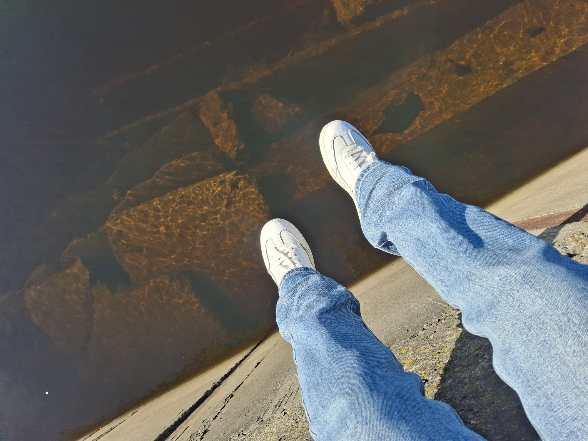 The image captures a first-person perspective, looking down at a pair of legs and feet positioned above a body of water. The person is wearing light blue, relaxed-fit jeans and clean white sneakers with white laces. The legs extend forward over an edge made of concrete or stone, which runs diagonally across the bottom right of the frame.
Beneath the person's feet is dark, somewhat murky water with a reddish-brown tint. The sunlight reflects strongly on the water's surface, creating shimmering, dappled patterns of light, particularly visible over the submerged structures. Below the surface, a series of large, dark, rectangular or block-like structures are visible. These appear to be either submerged steps, foundations, or retaining elements, suggesting a man-made construction underwater, possibly part of a dam, canal, or waterfront. The water's clarity allows for a good view of these structures, even with the light reflections.
The scene appears to be set outdoors on a sunny day, indicated by the bright highlights and shadows. The light conditions suggest it could be mid-morning or late afternoon. The concrete/stone edge where the person is situated shows some texture and variations in color, including patches of what might be moss or discoloration. There is no visible text in the image, and no specific emotions can be discerned. Given the location context of Chaikovsky, Russia, this could be a view over a river like the Kama or a reservoir.