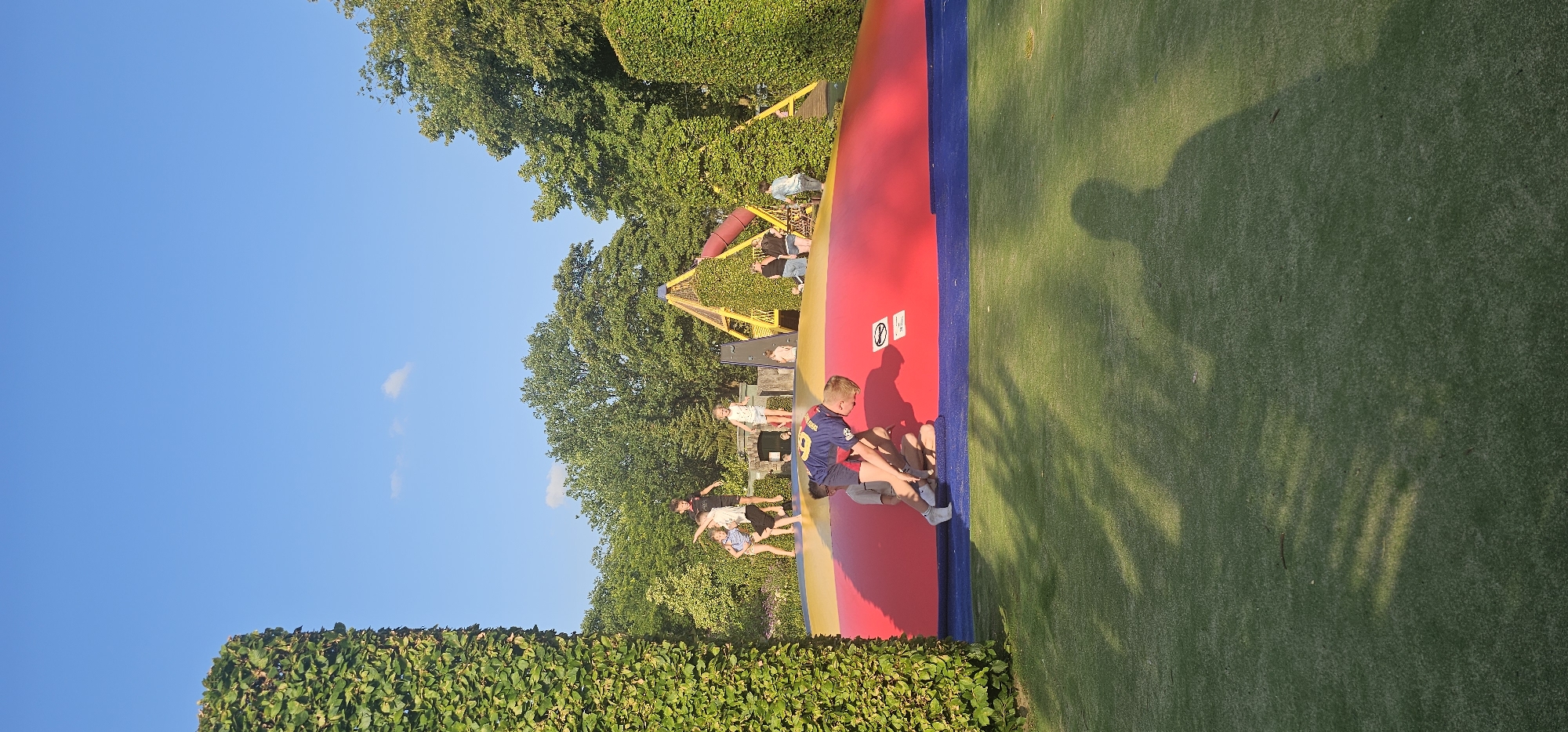 This image shows a vibrant outdoor playground scene in Groenlo, Netherlands, bathed in the warm glow of a sunny day. The focal point is a large, inflatable structure with sections in red, yellow, and blue, which appears to be a trampoline or bouncy castle. Several children are actively engaged on this inflatable surface, with some jumping and others interacting. In the foreground, a young boy in a blue and yellow jersey is leaning over, with another child sitting on the inflatable beneath him.
Behind the inflatable, a yellow, triangular climbing structure, possibly part of a larger play area, is visible, partially covered in greenery. The background is dominated by lush green trees and a clear blue sky. The ground surrounding the inflatable is covered in well-maintained green turf, with dappled sunlight and shadows creating a dynamic visual effect. Tall, manicured hedges frame the left and right sides of the scene, adding to the park-like atmosphere. The overall impression is one of joyful recreation and a bright, pleasant day spent outdoors.