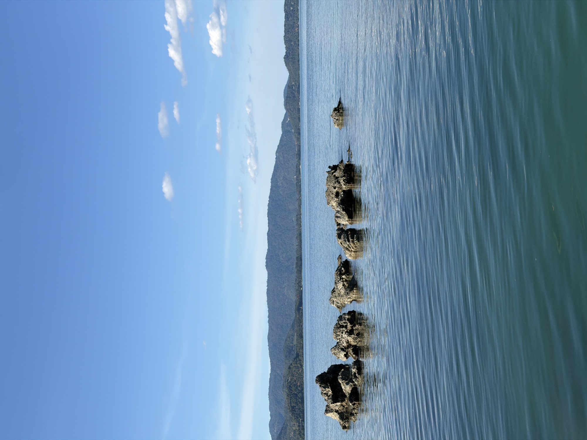 The image shows a tranquil seascape with a clear blue sky dotted with fluffy white clouds. The sea is calm and reflects the sky's blue, with gentle ripples disturbing the surface. A line of dark, rugged rocks emerges from the water, creating a natural barrier or perhaps a remnant of a past structure. The backdrop is a lush, green mountainous landscape that stretches along the horizon, hinting at a tropical or subtropical location. The sun appears to be high in the sky, suggesting it is daytime, likely midday or early afternoon, given the bright illumination and clear visibility. The overall scene conveys a sense of peace and natural beauty. The image is oriented vertically, with the sky on the left and the sea and land on the right.