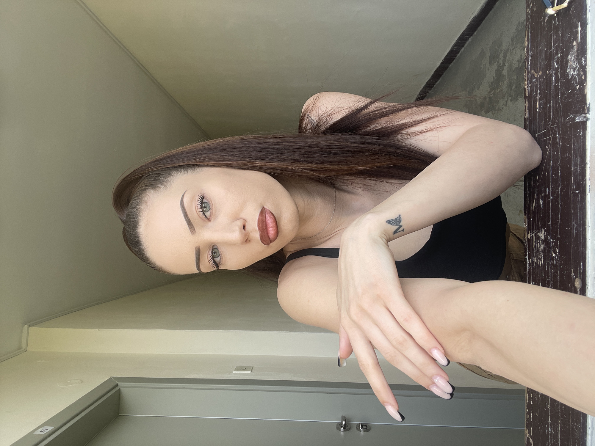 A young woman with long, dark brown hair, pulled back from her face, is prominently featured in a portrait-style shot. She has light-colored eyes, well-defined eyebrows, and is wearing reddish-brown lipstick. Her expression is calm and direct as she looks towards the camera. She wears a black tank top. A small black butterfly tattoo is visible on her left forearm. Her hands, positioned in the foreground, display long, manicured nails with a light pink base and black tips.
The setting appears to be an indoor or semi-enclosed area, possibly a hallway or stairwell within a building in Veliko Tarnovo, Bulgaria. The background includes plain, light-colored walls, with some trim visible. To the viewer's right, a section of dark, distressed wood or a doorframe shows significant wear, with scratches and peeling texture. To the viewer's left, a grey door or panel with a silver handle is partially visible. A small white label with the number "59" is affixed to this grey surface. The lighting is soft and natural, suggesting it is daytime.