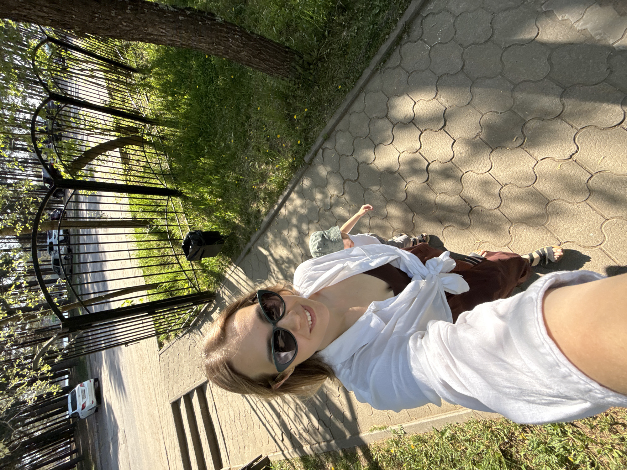 The image is a selfie of a woman with brown hair, wearing sunglasses and a white shirt tied at the front. She is smiling at the camera. In the background, there is a paved walkway bordered by grass and a black metal fence. Beyond the fence is a road with parked cars. A tree trunk is visible in the upper part of the image. Based on the information available, the location is Chaikovsky, Russia.