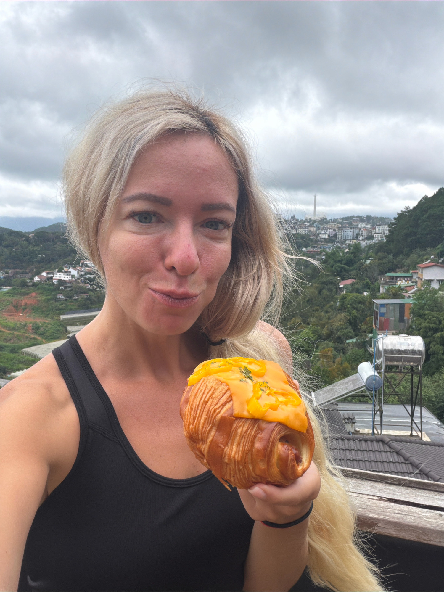 A blonde woman in a black athletic top is holding a croissant topped with melted cheese and herbs. She is smiling at the camera. In the background, a city with buildings and lush green hills is visible. The sky is overcast. The location appears to be a balcony or viewpoint in Da Lat, Vietnam, with a water heater and tiled roofs in the foreground. A distant tower can be seen in the cityscape.