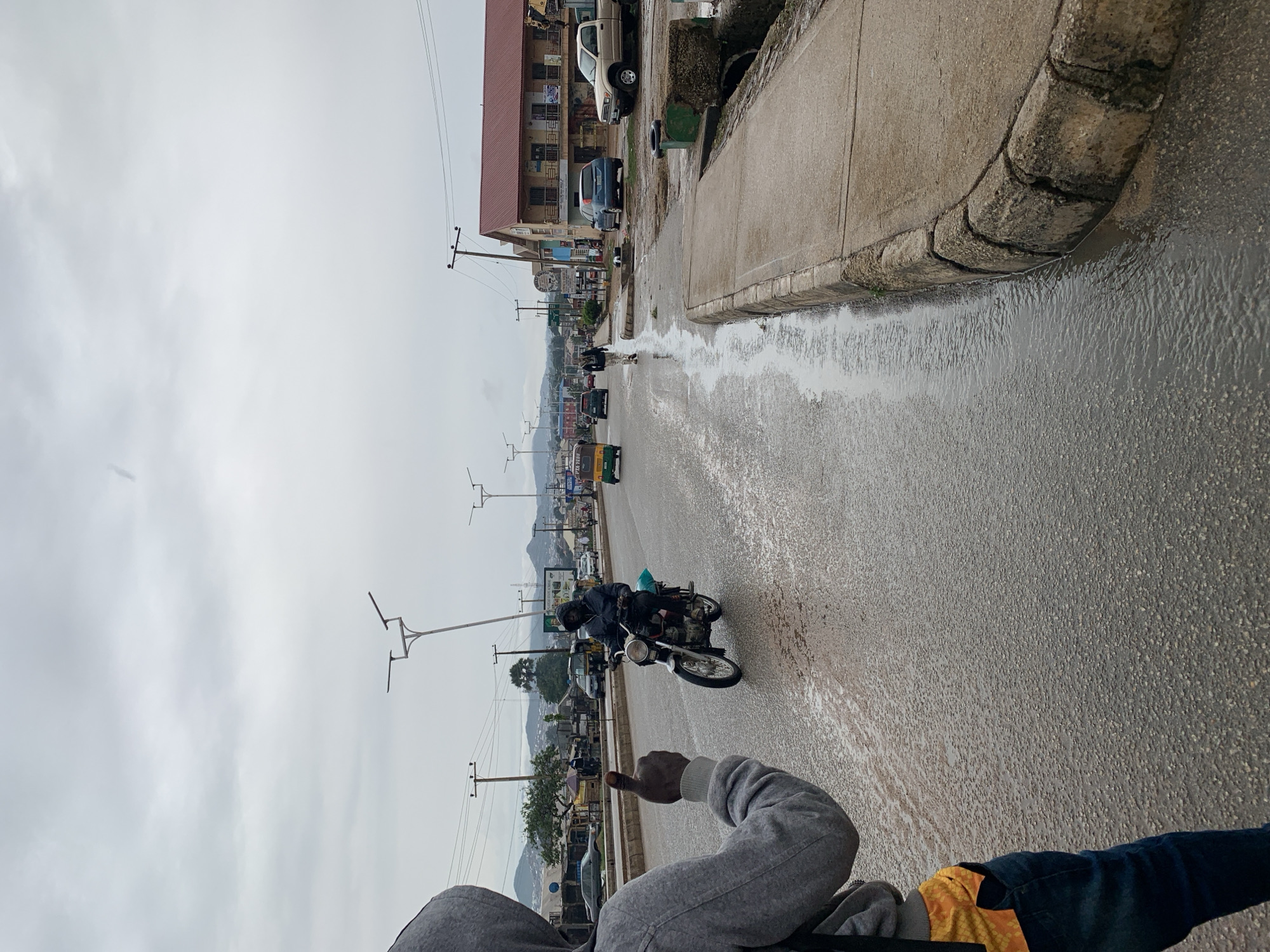 The image, currently oriented sideways, captures a street scene on an overcast day, with a wet road indicating recent rainfall or standing water. In the immediate foreground, a person wearing a grey hoodie and dark trousers has their arm extended, giving a clear thumbs-up gesture, suggesting approval or acknowledgement.
In the mid-ground, a person wearing a dark helmet and jacket is riding a motorcycle on the wet asphalt road, moving away from the viewer towards the right side of the frame. The road surface is dappled with water and reflections. To the right of the road, a concrete sidewalk with a rounded curb separates the pedestrian area from the street, with a visible flow of water in the gutter.
Along the left side of the street, several multi-story buildings, some appearing commercial, line the roadside. Various vehicles, including cars and a distinctive green and yellow three-wheeled vehicle (likely a keke Napep or tuk-tuk), are present on the road and parked along its edges, extending into the distance. Numerous utility poles with overhead power lines are spaced along the street. In the far background, low-lying hills or mountains are visible beneath the uniformly grey, cloudy sky. There is some faint, illegible text on distant signs attached to buildings. The scene conveys a moment of daily life in an urban or semi-urban setting, likely after a period of rain.