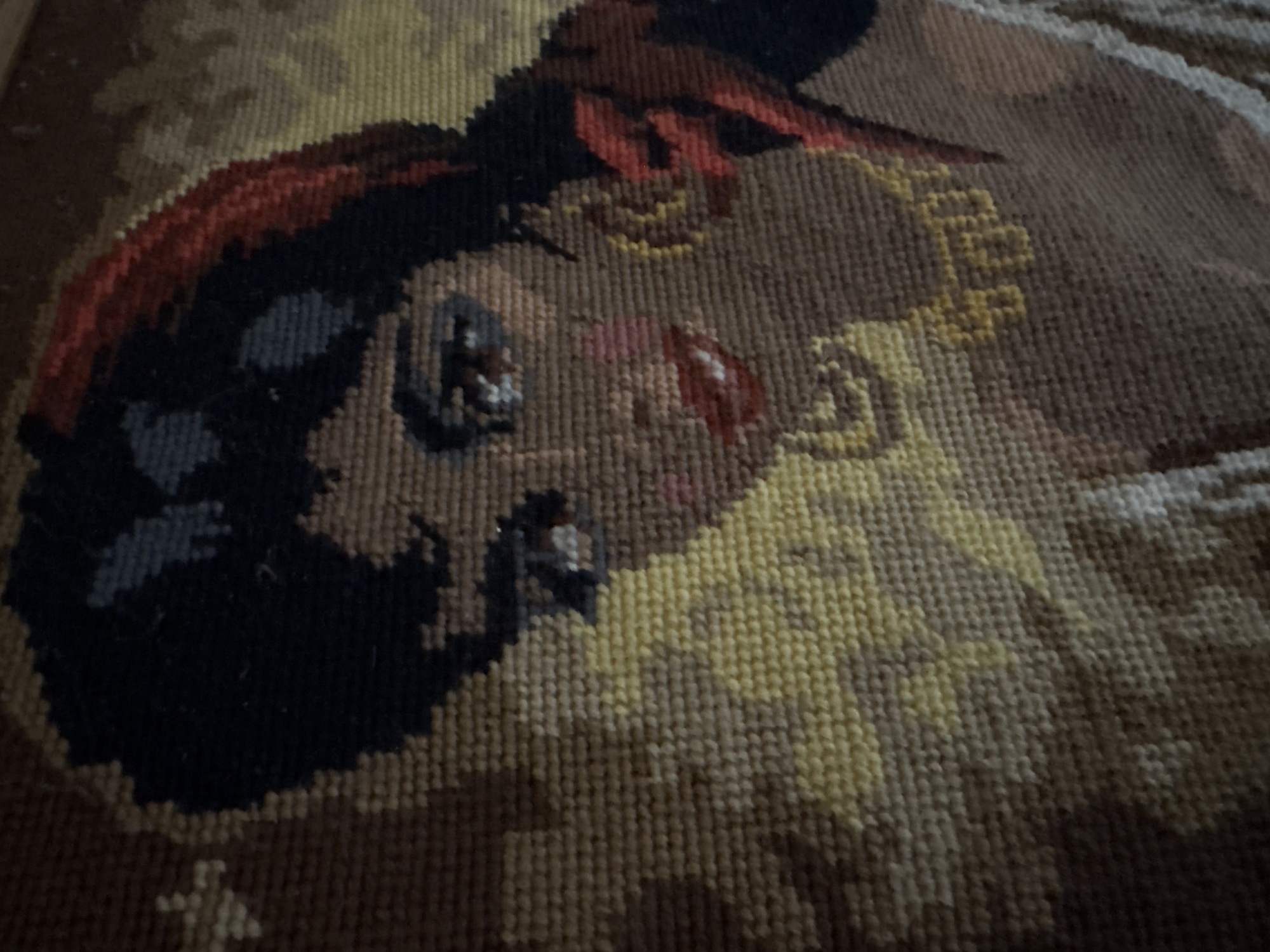 This is a close-up, head-and-shoulders, needlepoint tapestry depicting a woman with dark hair styled in a bouffant and a red headscarf. She has dark eyes, prominent eyebrows, and her lips are painted red. She wears large, gold hoop earrings and a gold necklace. Her attire appears to be a white garment. The background is textured in shades of brown and yellow. The image's location in Bellheim, Germany is not determinable from the visual information provided.