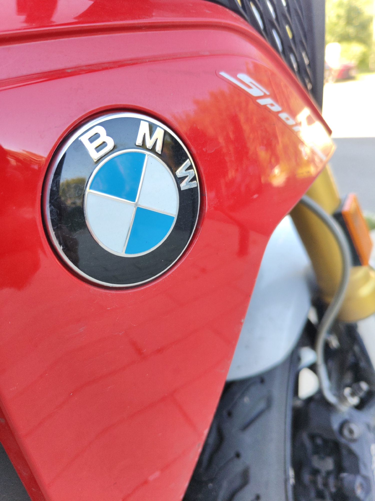 A close-up, eye-level, outdoor shot shows the red bodywork of a BMW motorcycle, prominently featuring its round, chrome-rimmed emblem with a blue and white checkered design and the letters "BMW" in white text. The word "Sport" is visible in white lettering on the red fairing to the right of the emblem. The background is blurred but suggests a sunny day with some greenery and possibly other vehicles. The motorcycle appears to be parked, based on the lack of motion blur. The location is identified as Kommunarka, Russia.