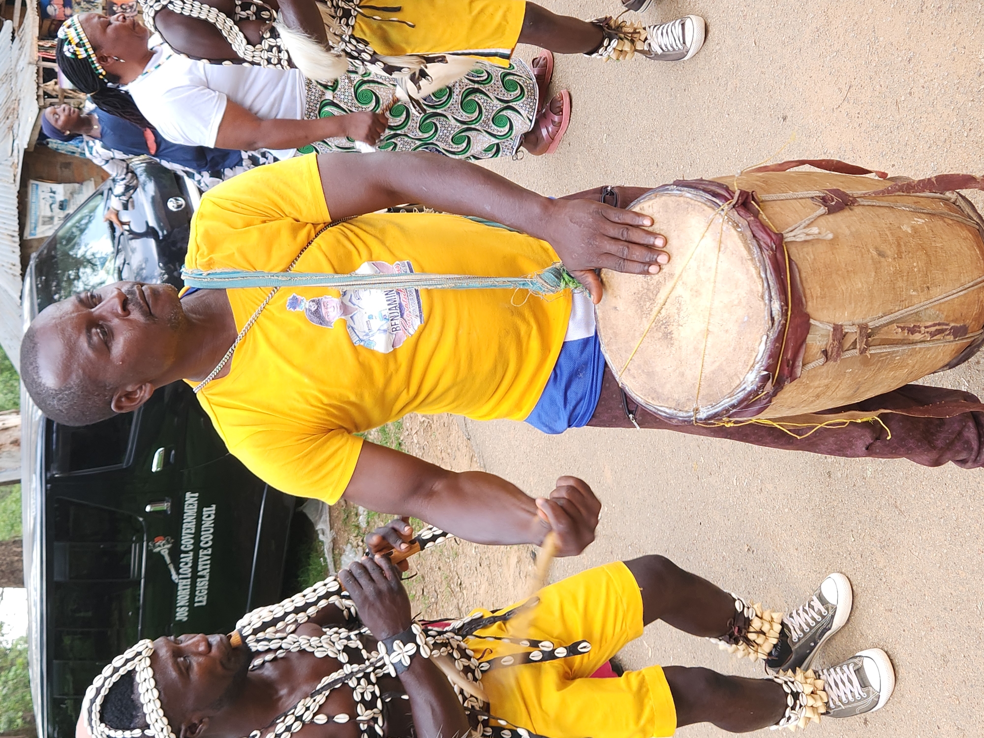 In Jos, Nigeria, two men are participating in a cultural event. The man in the foreground, wearing a yellow t-shirt with a picture of a uniformed individual on it, is playing a large, traditional drum. He is also adorned with a beaded necklace and a blue lanyard around his neck. The other man, on the left, is dressed in traditional attire, including a headdress decorated with shells, a beaded necklace, and yellow shorts. He appears to be playing a wind instrument. Behind them, a black van with "JOS NORTH LOCAL GOVERNMENT LEGISLATIVE COUNCIL" written on its side is parked. Several other people, including women in traditional clothing, are visible in the background. The event is taking place outdoors on a dirt or gravel surface, possibly a street or open area.