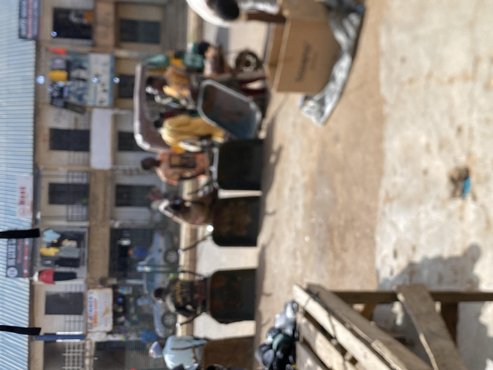 The image shows a street in Bukuru, Nigeria. There are multiple rickshaws lined up on the right side of the street. People are sitting on them. Buildings are visible on the left side of the street. Some have signs with writing on them. There is some equipment or furniture on the right.