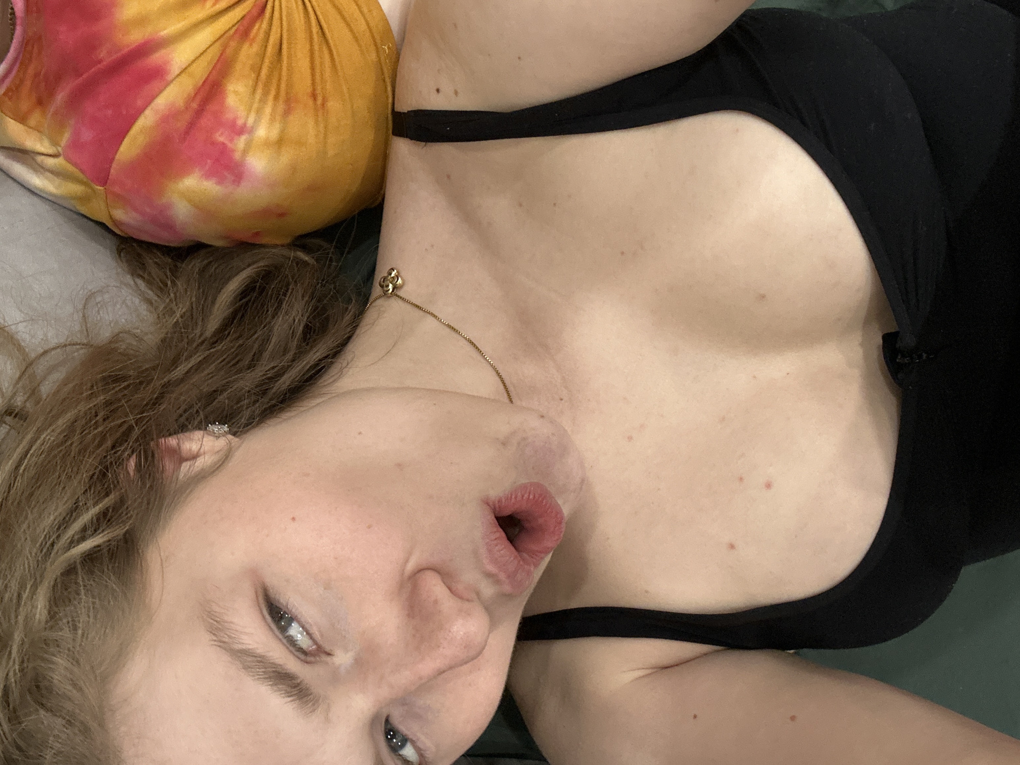 A woman with fair skin and light brown hair is lying down, facing the camera. She is wearing a black tank top and a gold necklace with a clover-shaped pendant. Her mouth is slightly open. She appears to be resting on a pillow with a tie-dye pattern. The environment appears to be indoors, but the precise location is not determinable based on the given image. There is no indication that the image was taken in Kazan, Russia.