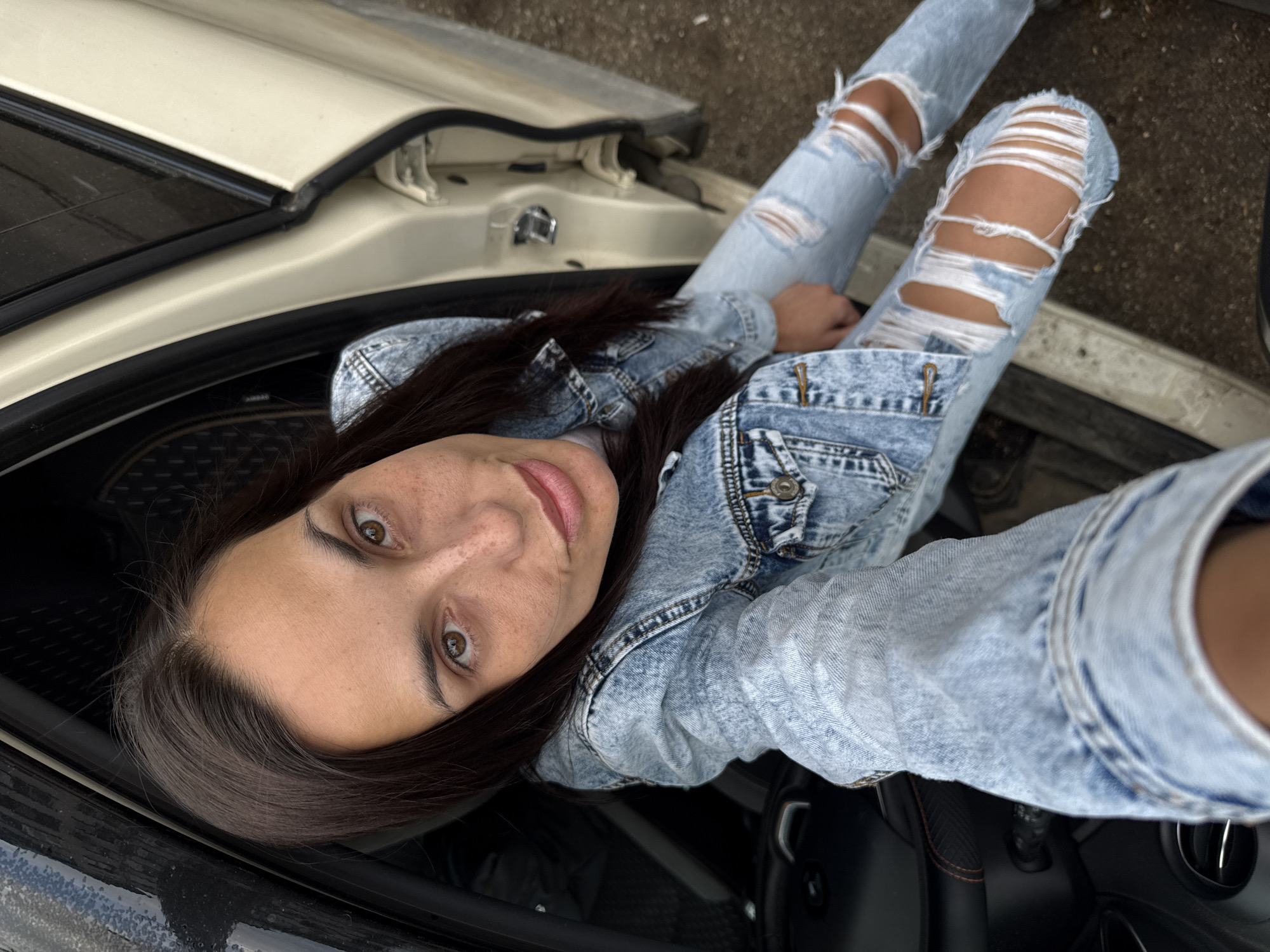 A woman is sitting in a car, with her legs extended out the window, wearing ripped jeans and a denim jacket. The car door is open, revealing a cream-colored interior trim. The car is parked on a street in Pleven, Bulgaria. The background is an overcast sky. The woman is looking directly at the camera.