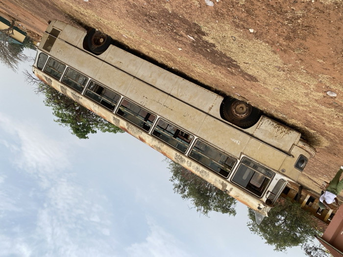 The scene, located in Ayangba, Nigeria, presents an outdoor environment under a light blue sky with scattered white clouds, viewed with an upward diagonal tilt. The ground consists of reddish-brown earth with patches of dry, light-colored vegetation and scattered white debris. A prominent, heavily weathered, light-colored (beige/off-white) bus is centrally positioned, appearing abandoned and partially sunken into the ground. The bus displays significant rust, peeling paint, and dirt accumulation on its exterior and roof. Most windows are missing or broken, exposing empty frames. Faint dark lettering, possibly "T.S.BOL," is visible on the side panel near the front. The bus's wheels are dark and appear to be either missing tires or severely deflated, submerged in the soil. One individual, wearing a white short-sleeved top and green trousers, stands on the far left of the frame near the front of the bus, facing left. In the background, a reddish-brown building with a dark roof is visible on the left, with additional smaller, lighter-colored buildings with dark roofs partially seen on the right. Trees with varying degrees of foliage are present in the mid-ground and background.