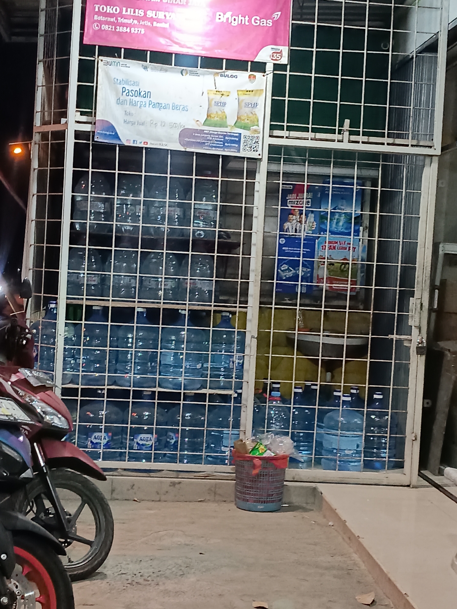 The image shows a storefront in Jetis, Indonesia, likely a small convenience store or warung. The storefront features a metal grate structure that displays various products, primarily large bottles of drinking water. The bottles are stacked on shelves within the grate. Above the grate, there are several signs. One sign at the top is pink and displays text in what appears to be Indonesian, with the words "TOKO LILIS SURYA" and "Bright Gas" prominently visible. Below this, another white sign has the BUMN logo and text related to "Stabilisasi Pasokan dan Harga Pangan Beras" (Stabilization of Supply and Price of Rice). Further down, more signs are visible, including one for "MIZONE" and others advertising beverages. In front of the storefront, two motorcycles are parked. The ground in front of the store is paved, and there is a red and blue striped trash can filled with plastic bags and wrappers. The overall scene suggests a retail environment selling food and beverages, possibly with a focus on essential goods like rice.