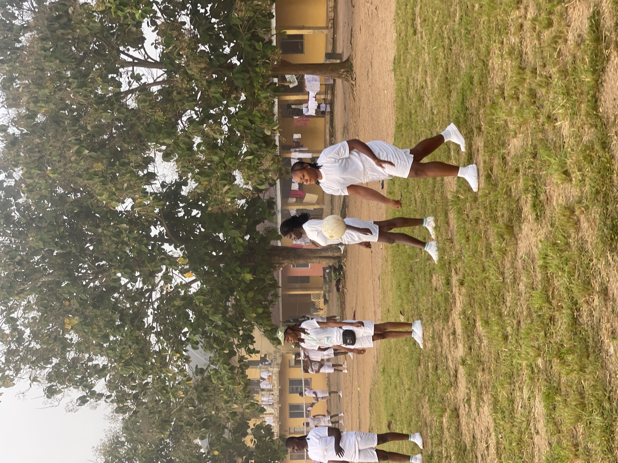 In Ise - Ekiti, Nigeria, a group of young women in white t-shirts and white shorts are engaged in an outdoor activity on a grassy field with dirt patches. The women are wearing white sneakers, and some have green and white striped socks. One woman in the foreground on the right is holding a light-colored ball. In the background, there are yellow and pink buildings with some laundry hanging outside. Several trees with green leaves are present on the field and in front of the buildings. The sky is overcast and appears hazy. A volleyball net is visible in the mid-ground.